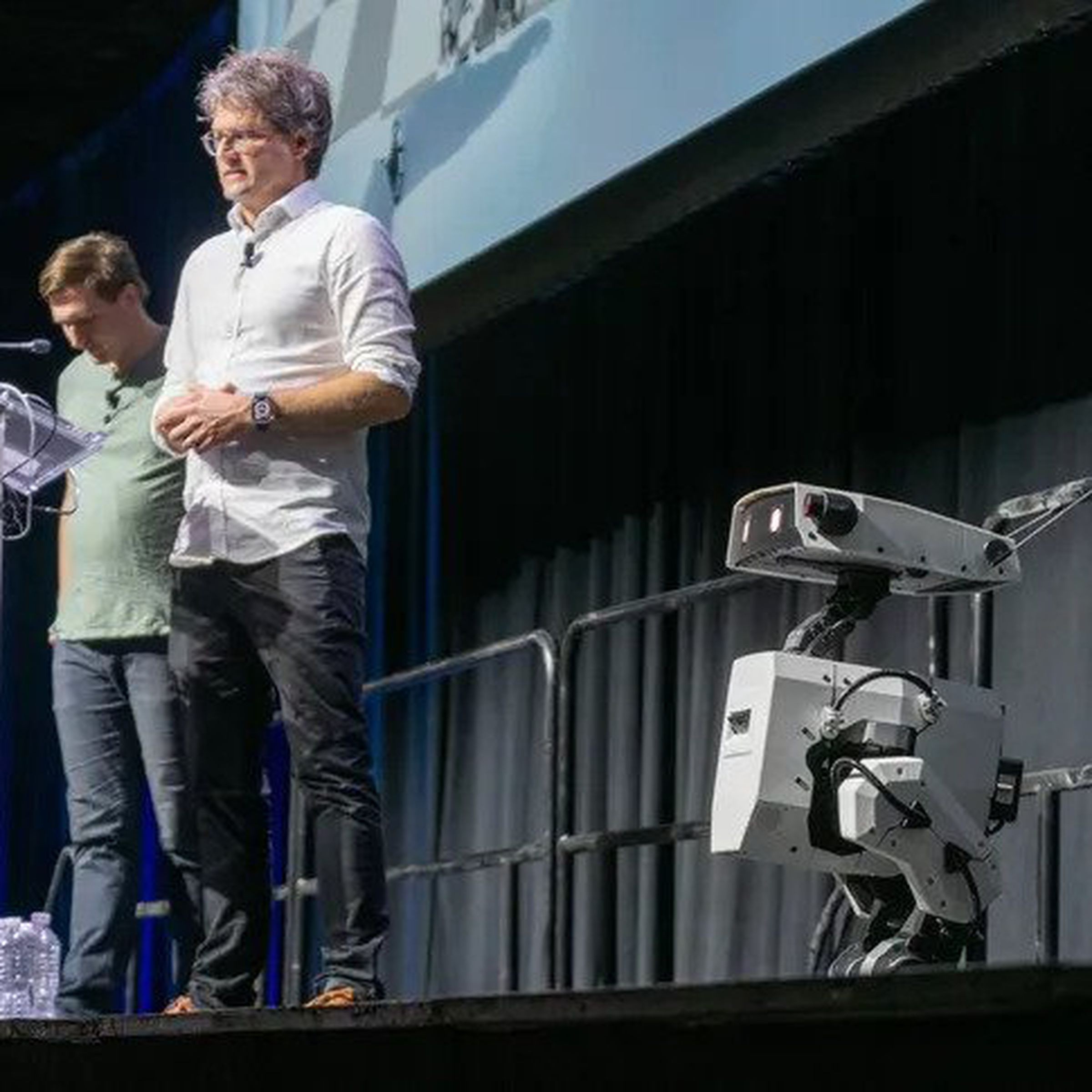 Two men appear in the background presenting an adorable bipedal concept robot, which stands in the foreground.