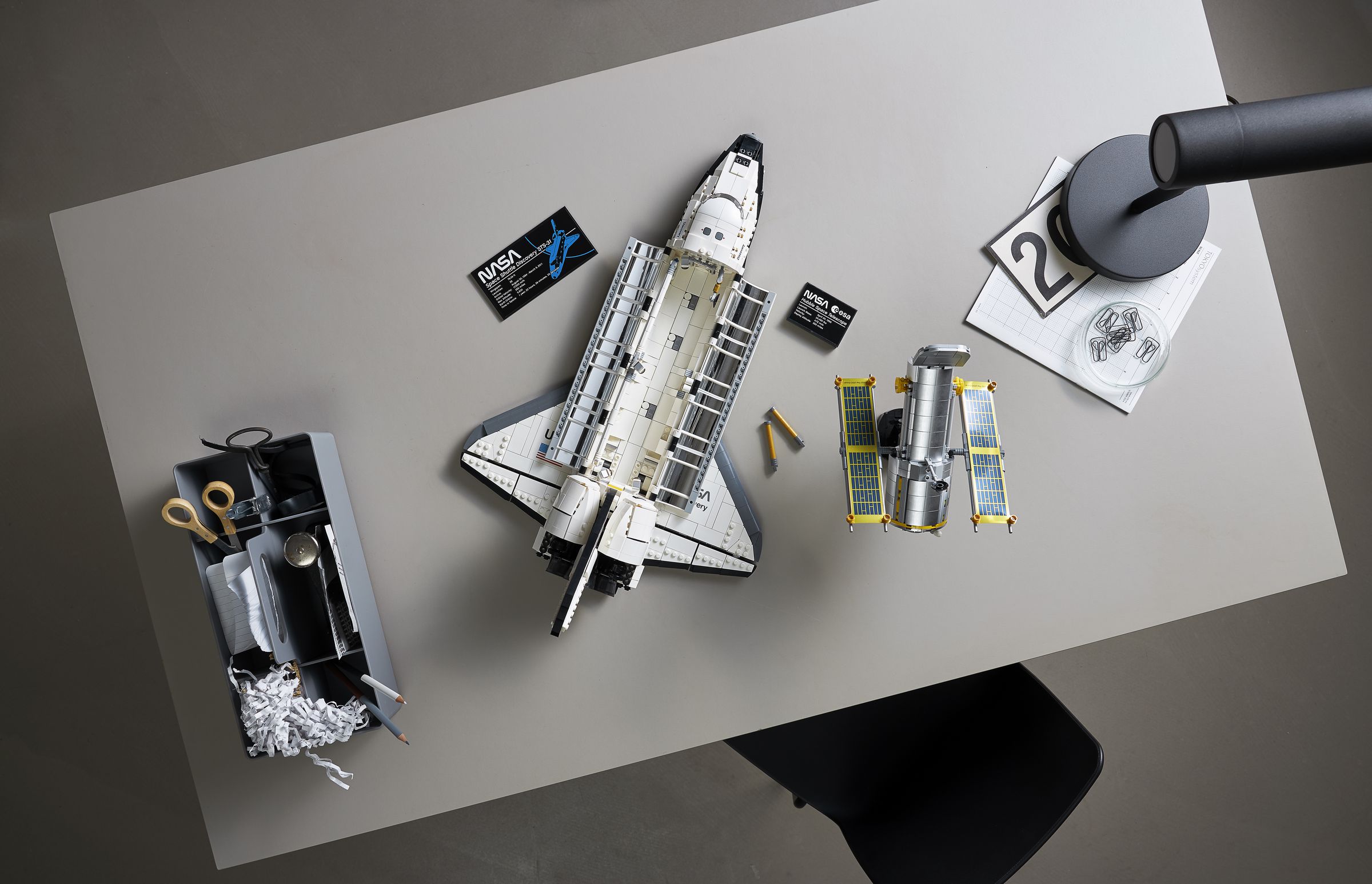 A bird’s eye view of the Lego space shuttle on a table, with it’s payload bay open to show the shiny interior of its doors. The Hubble telescope is next to the shuttle.