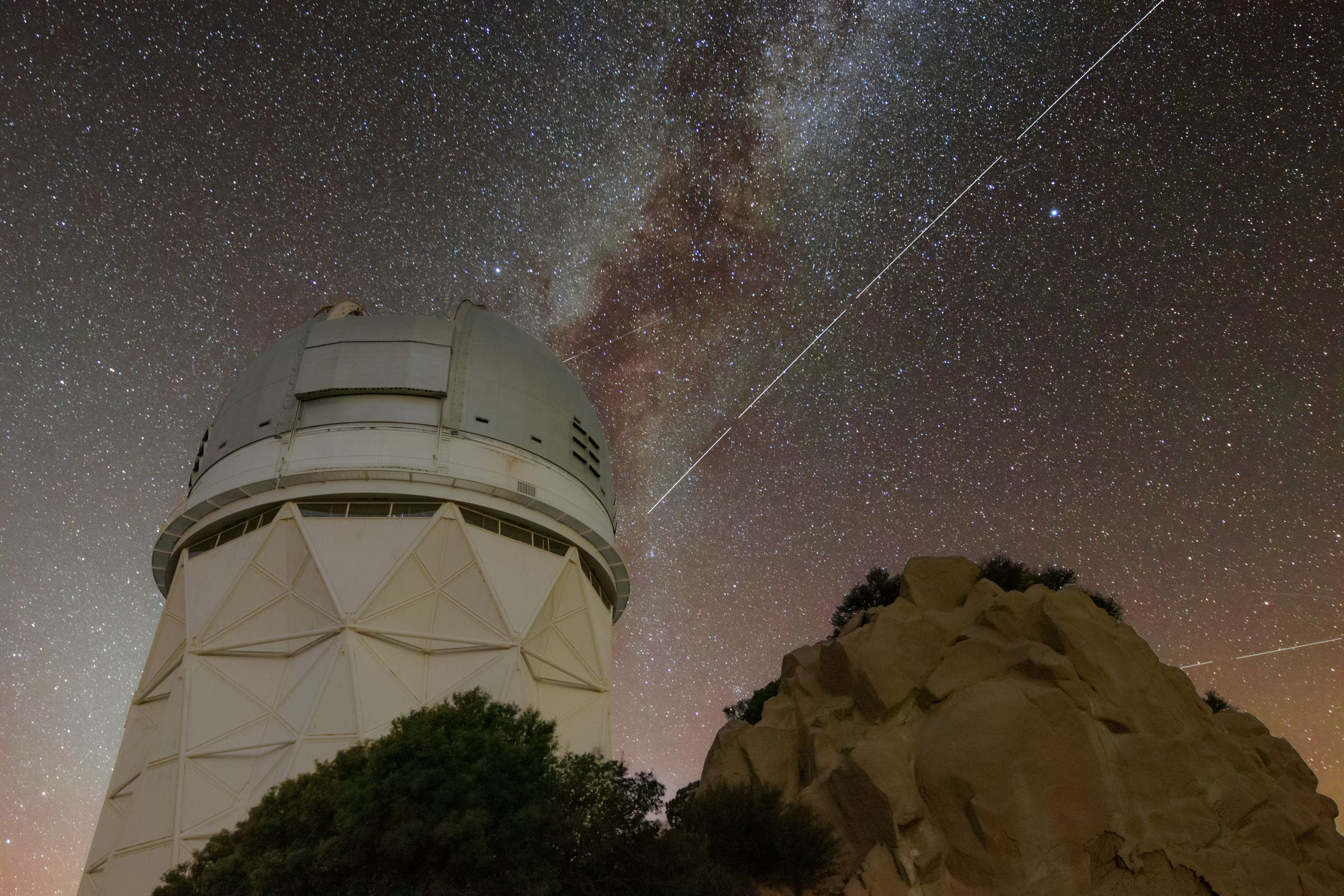 Bright white streaks across a glorious night sky.  A domed telescope and rock dominate the bottom of the photo.