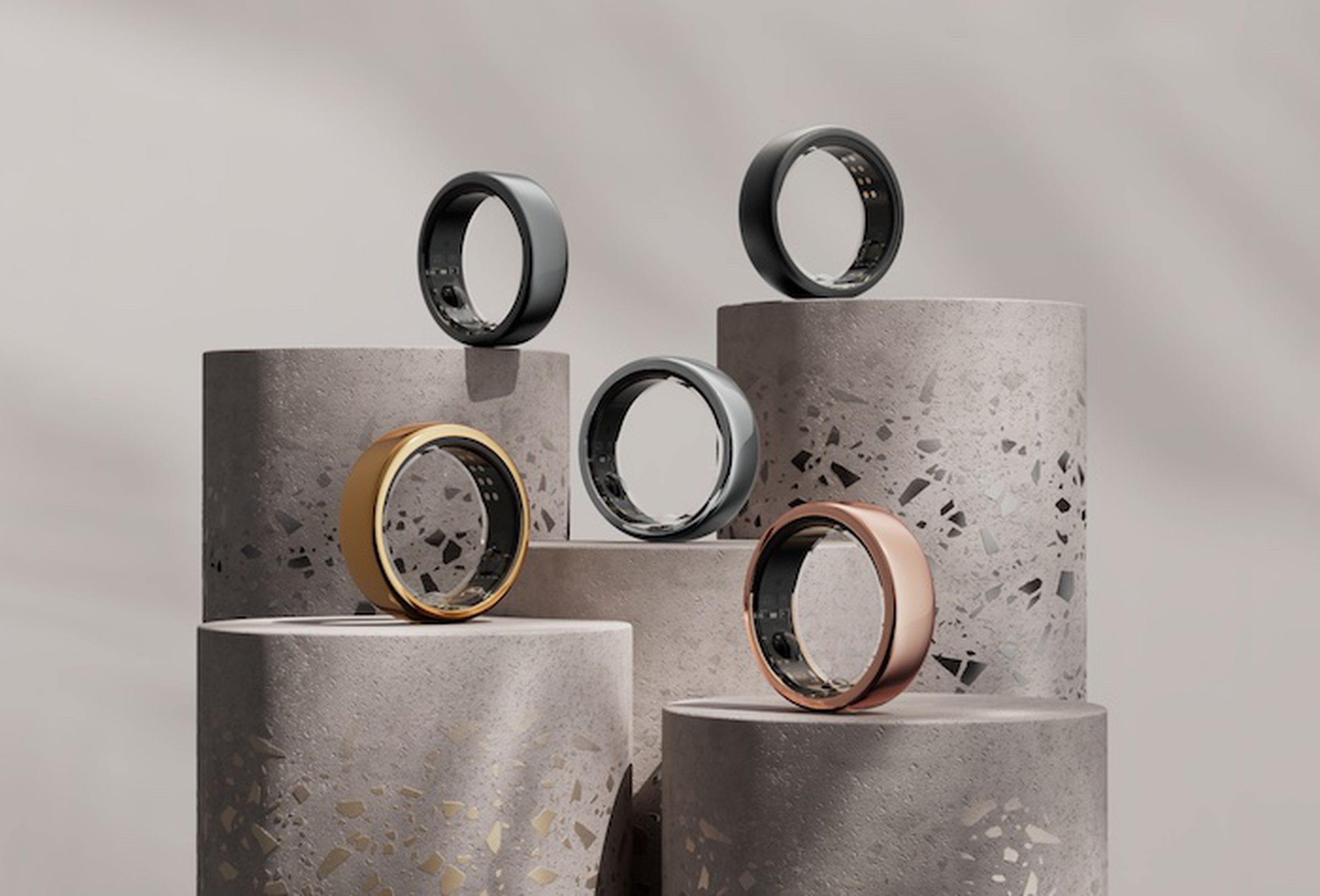 A collection of five Oura smart rings are arranged on concrete pedestals of varying heights. There’s a glossy gold ring, a rosy gold ring, a silver one in the center, a gunmetal gray one, and a matte black one.