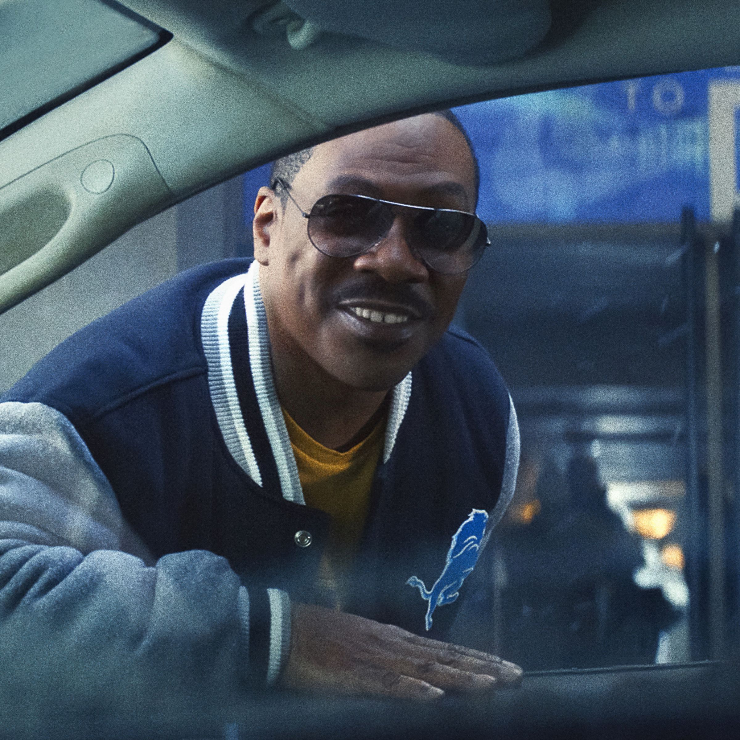 A man wearing aviators and a letterman jacket leaning over to peer into the window of a parked car.