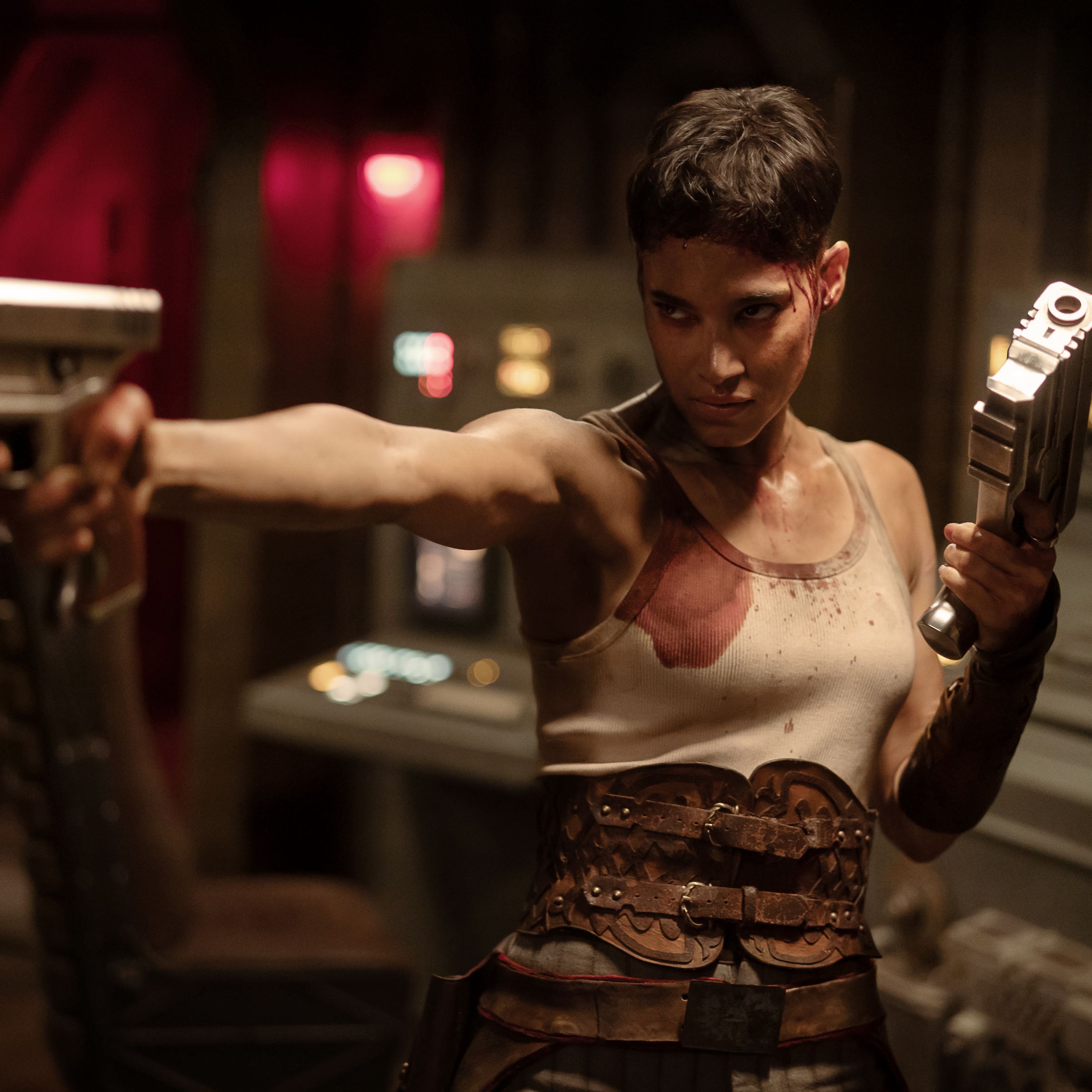 A woman in a tank top, a combat girdle, and pants holding two guns while standing in some sort of control room.