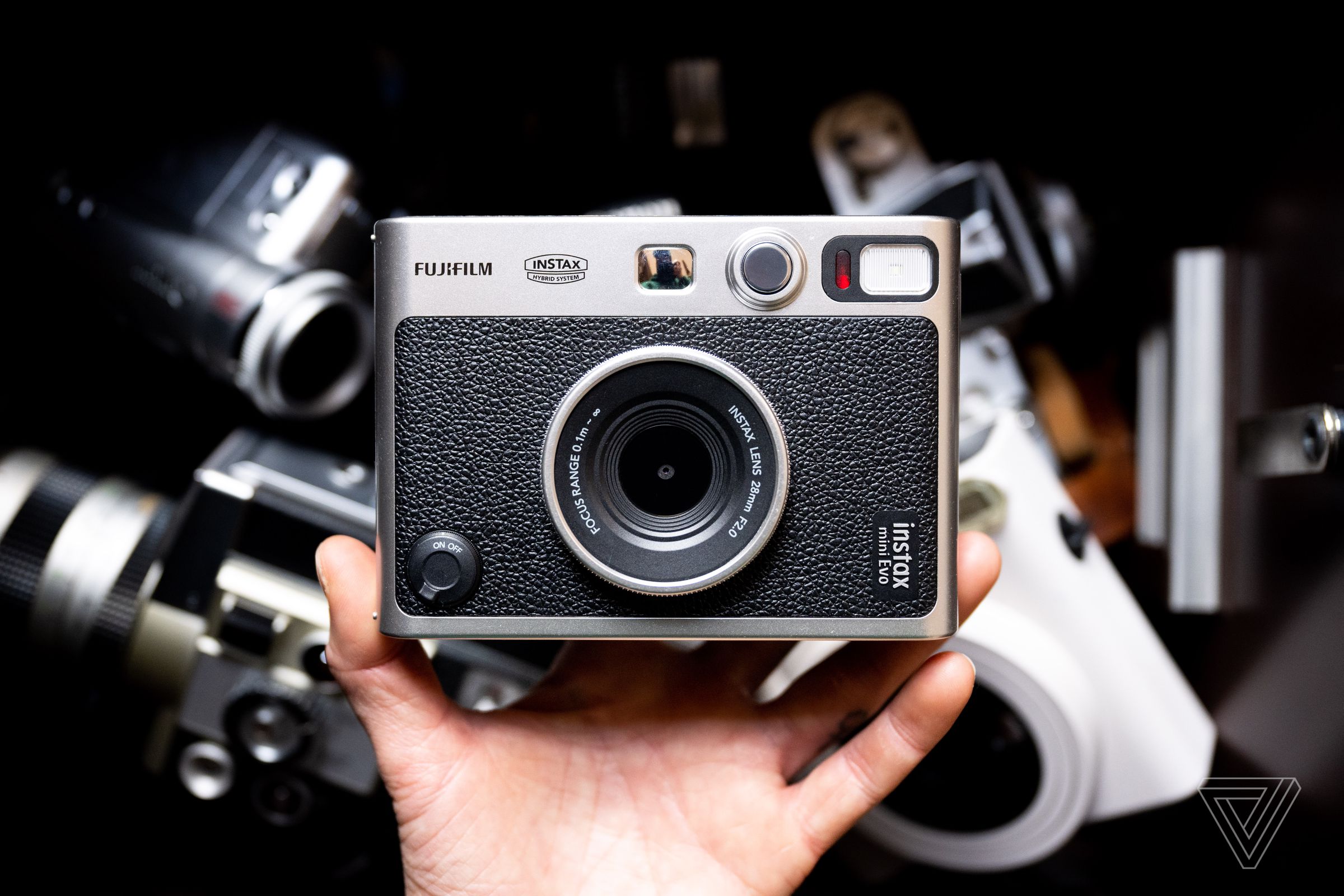 The Instax Mini Evo being held up with a hand against a dark background with cameras that are out of focus.