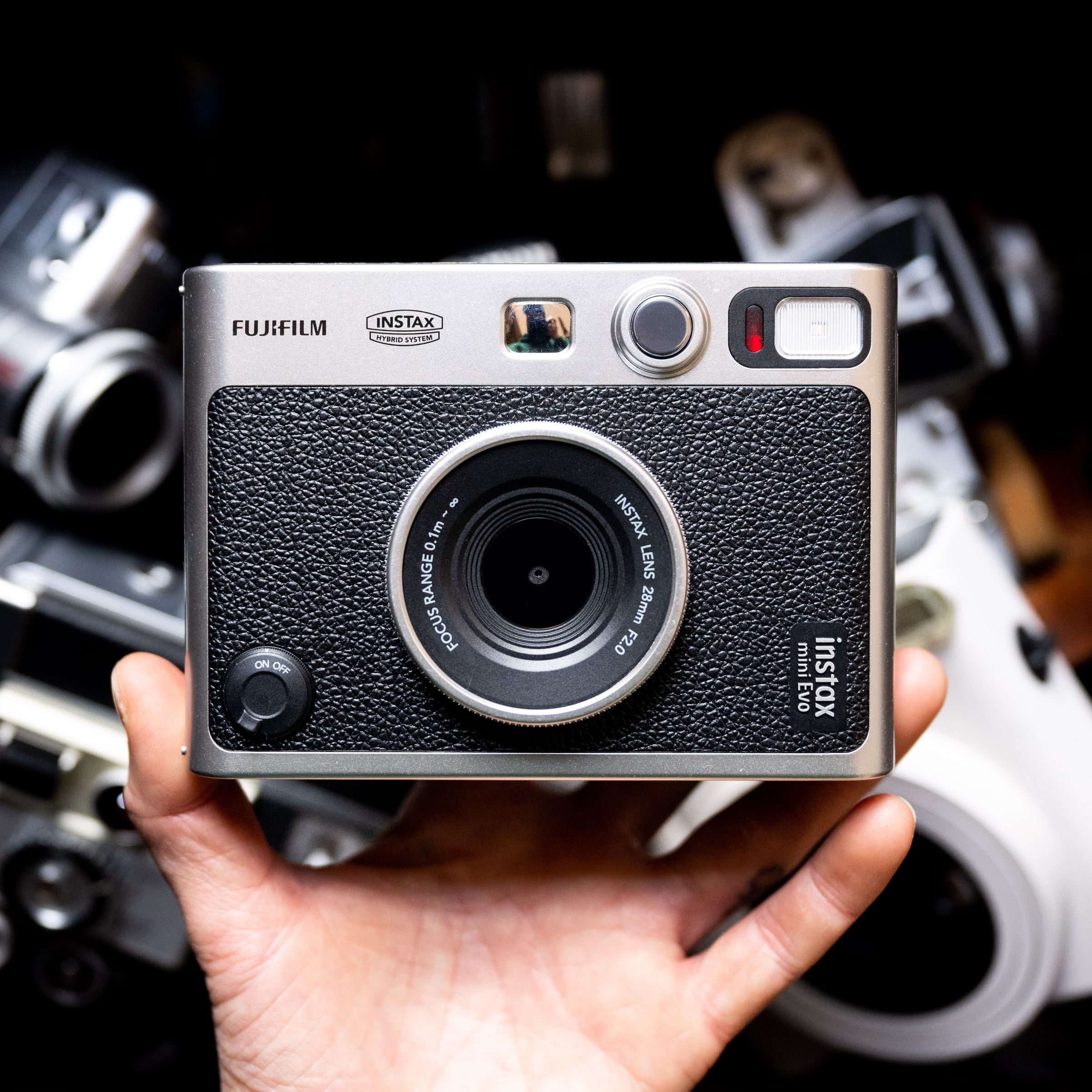 The Instax Mini Evo being held up with a hand against a dark background with cameras that are out of focus.