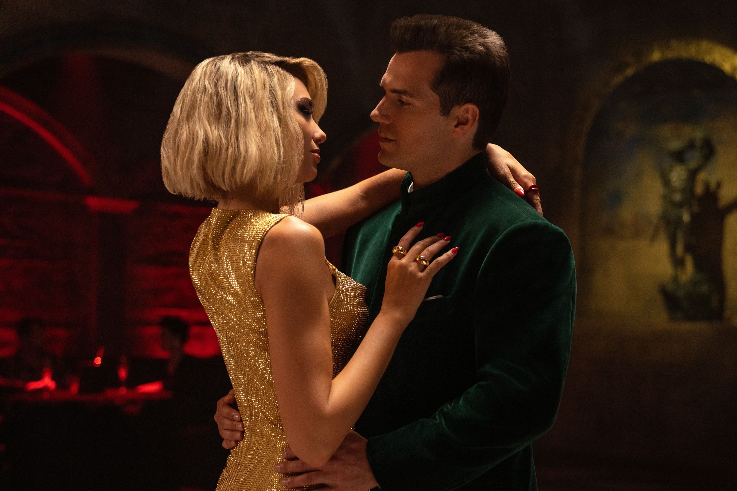 A blonde woman in a shimmering golden dress dancing closely with a brown-haired man wearing a green crushed velvet suit.
