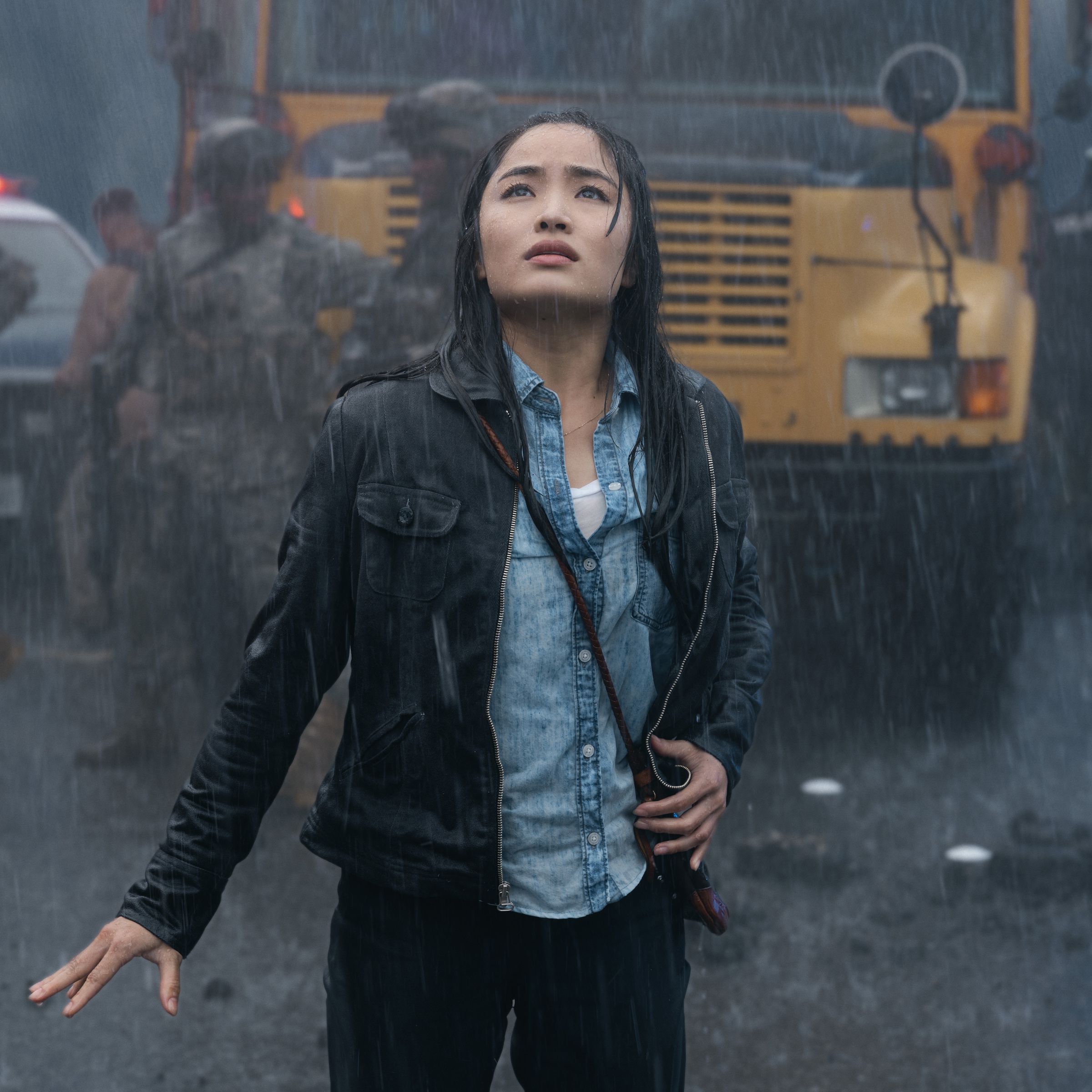 A woman in a blue shirt, black pants, and a black jacket standing in the rain in front of a school bus as she looks up in dismay at something towering above her.