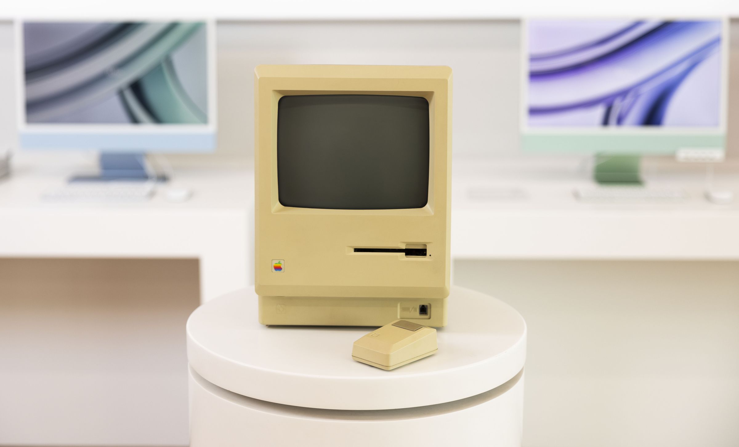 An original Macintosh sits on a pedestal in a brightly lit room with two Apple silicon iMacs behind it in blue and green.
