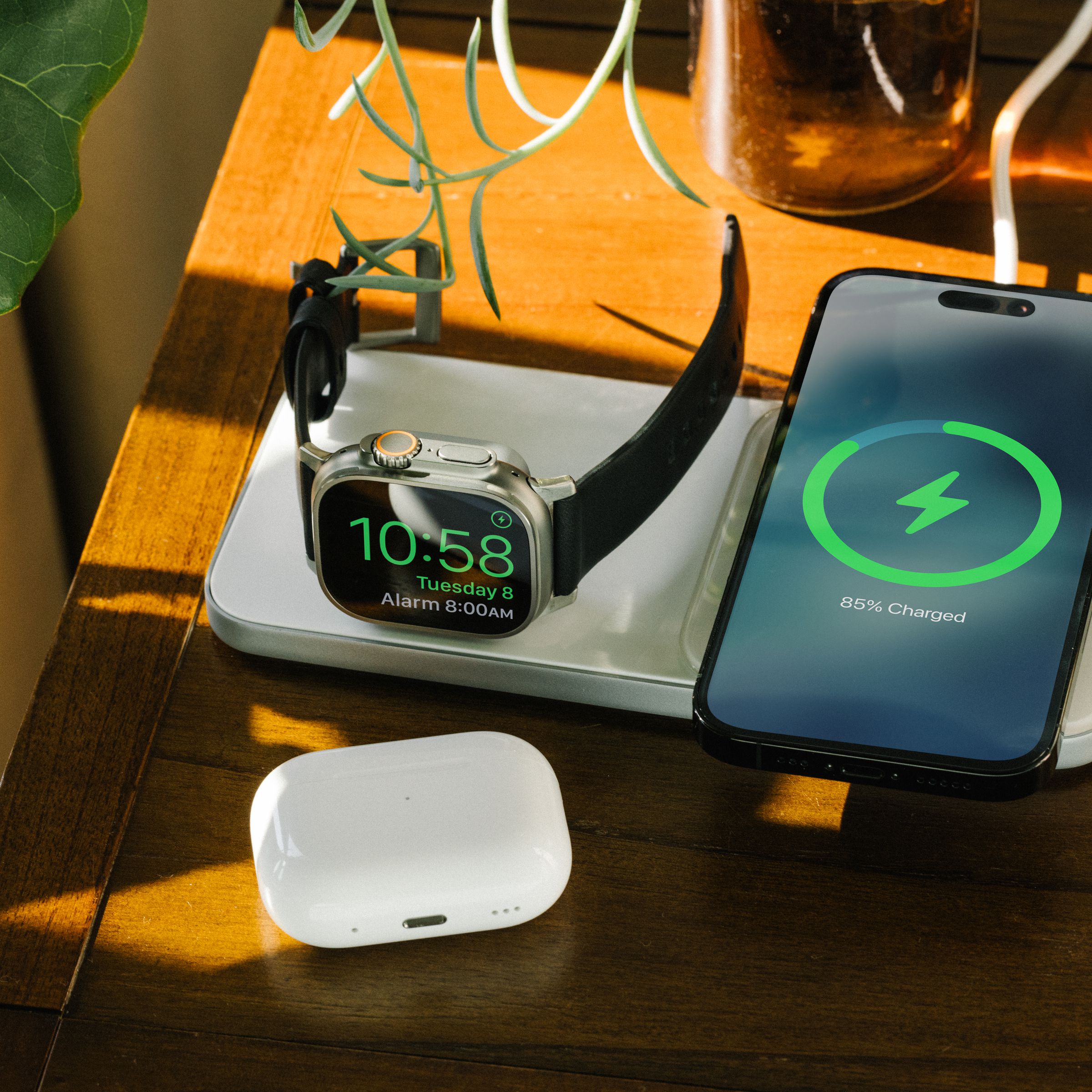 An iPhone and Apple Watch on a table charging on Nomad’s Base One Max wireless charger, with a set of AirPods near.