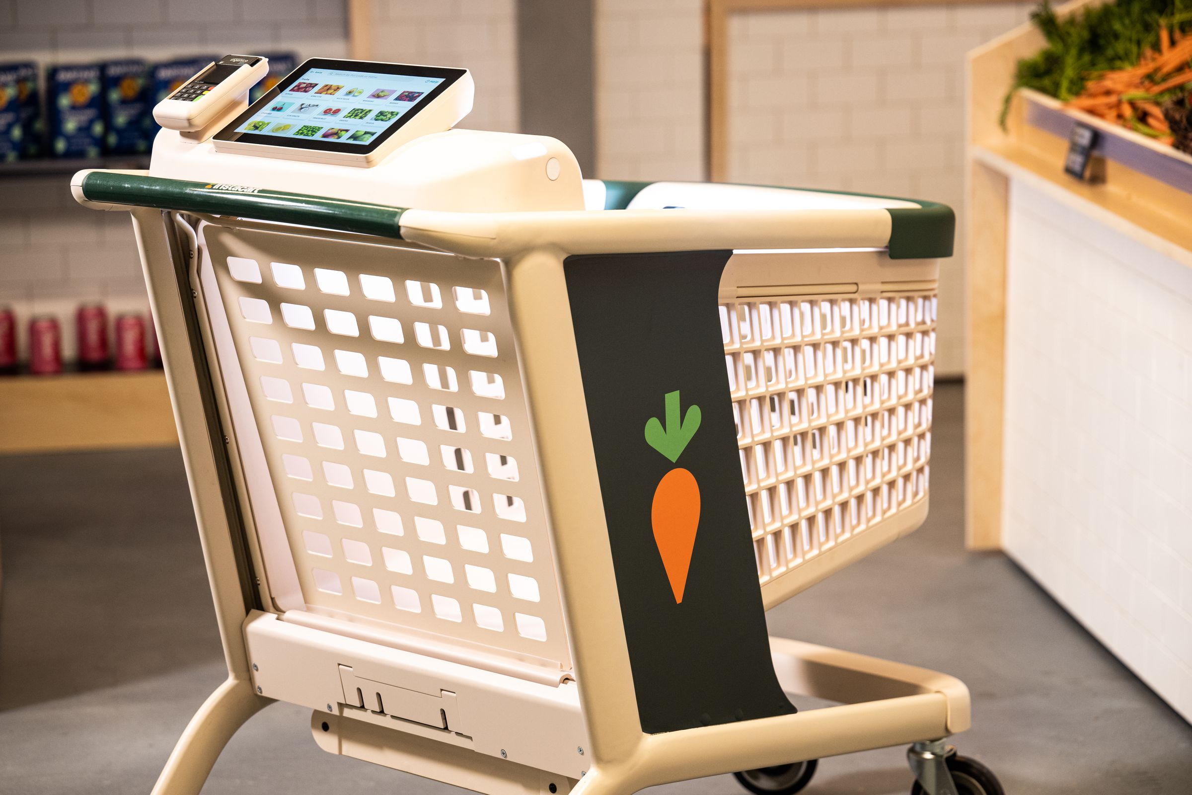 In the middle of a modern grocery store, with smooth concrete flooring, white oak shelving and white tiled walls, there’s a beige and thick-bodied plastic shopping cart. The cart has a big orange and green carrot logo printed on the side, and a tablet computer affixed to the top where a child would normally sit, as well as a digital payment terminal.