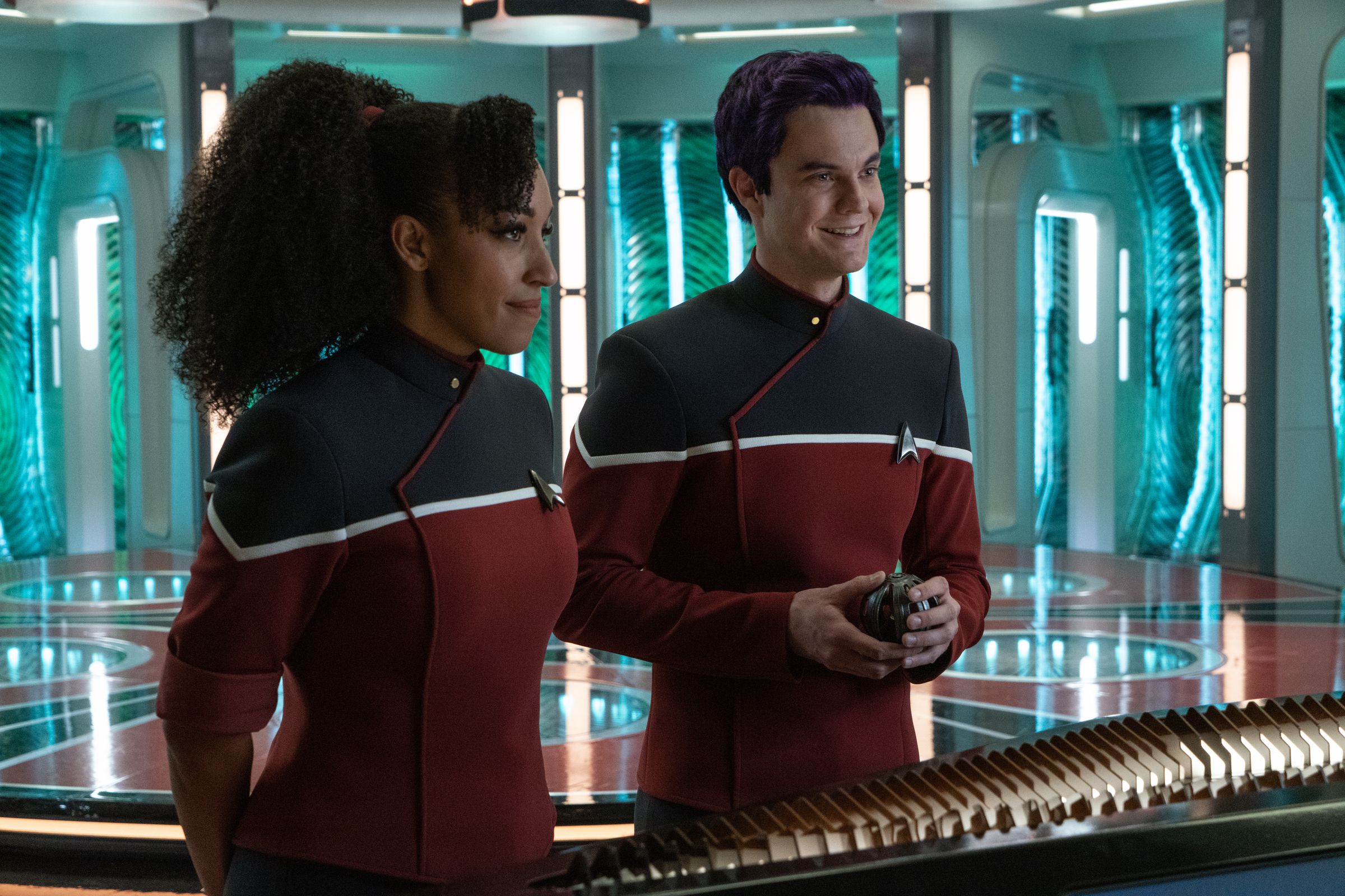 An image of a Black woman with her hair in a ponytail and a white man, with purple hair. They are standing in the transporter room of a Star Trek set and wearing black and red uniforms.