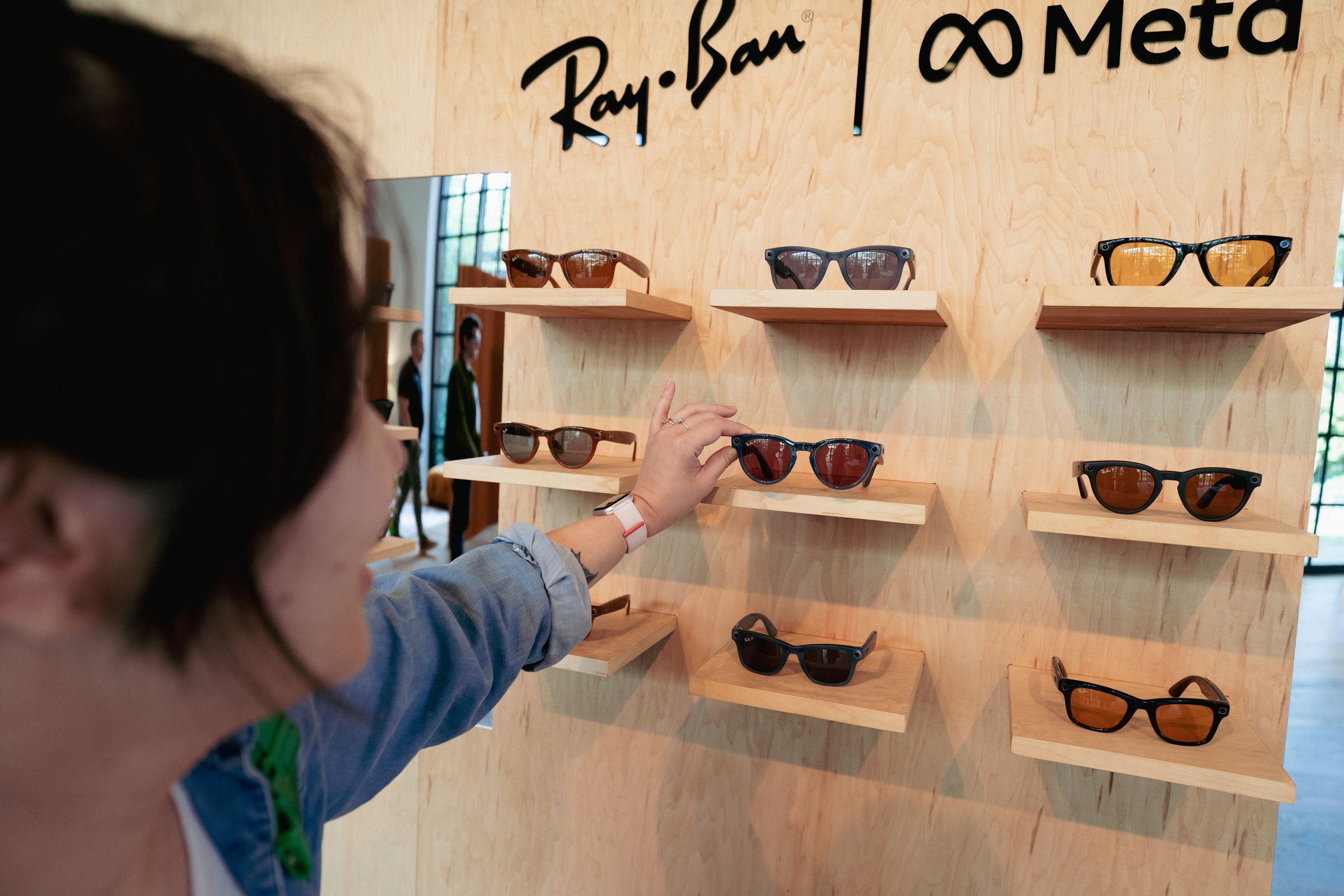 Woman reaching for blue pair of Ray-Ban Meta Smart Glasses from a display case