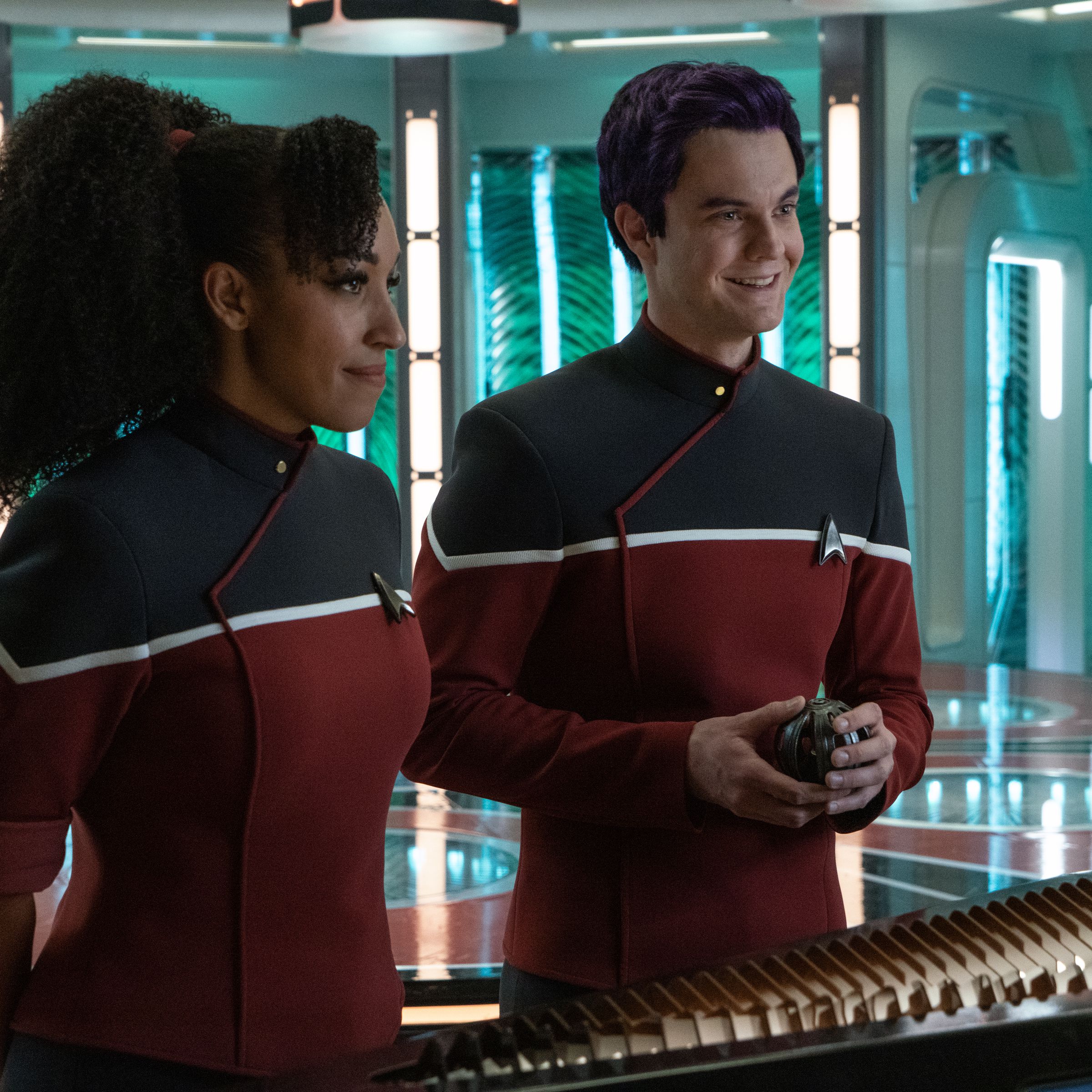 An image of a Black woman with her hair in a ponytail and a white man, with purple hair. They are standing in the transporter room of a Star Trek set and wearing black and red uniforms.