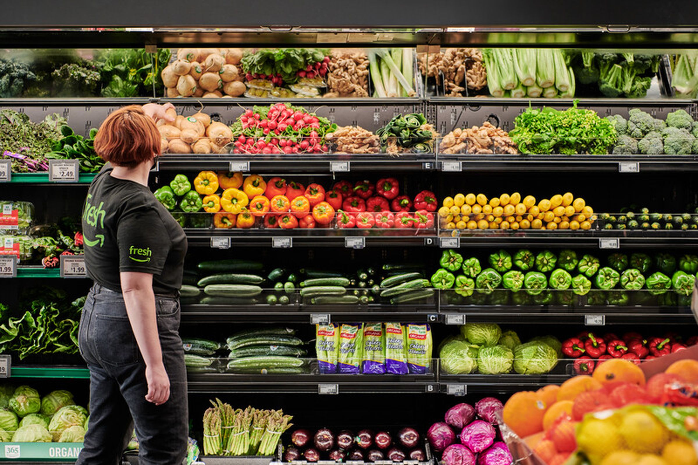 A person wearing an Amazon Fresh shirt browsing produce at a grocery store.