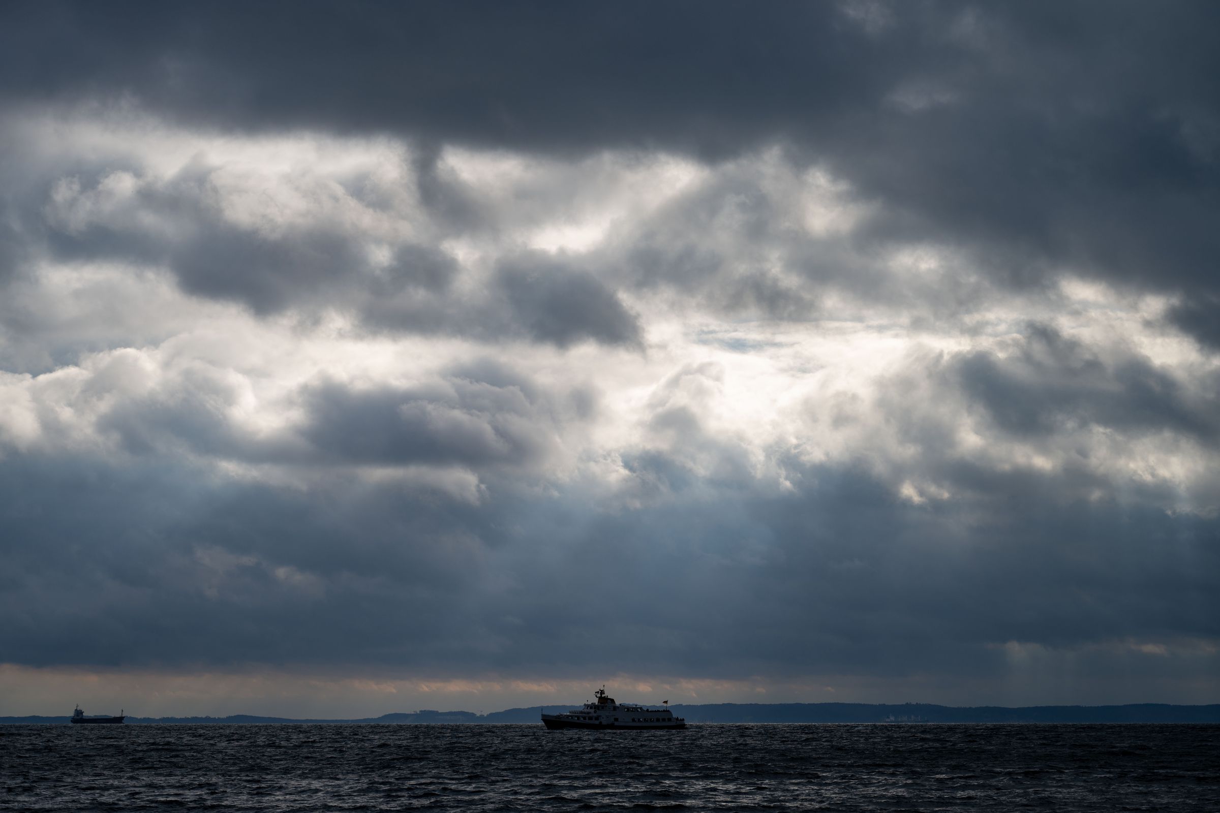 A ship on the sea is seen in the center of the photo. Above it, rays of sunlight pierce through gaps in thick clouds.