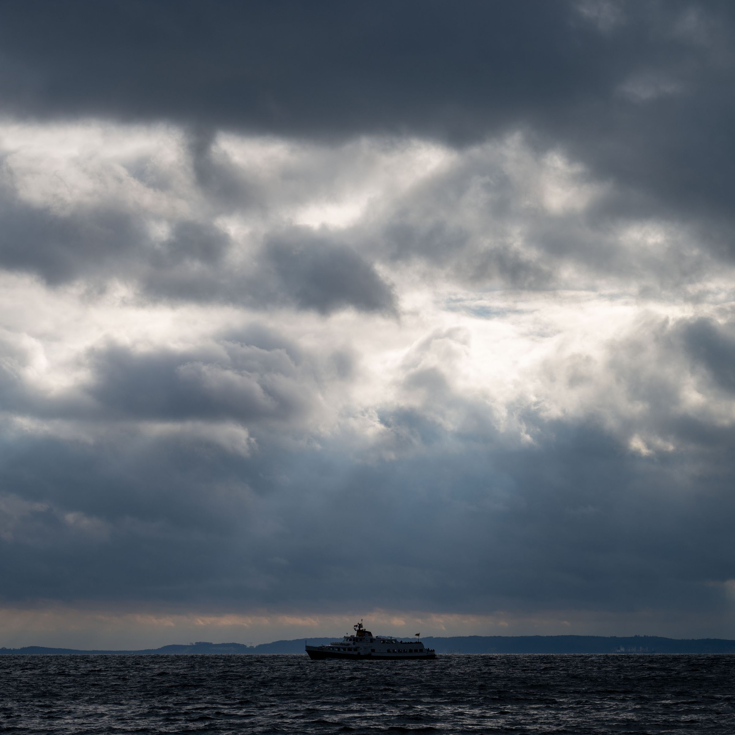 A ship on the sea is seen in the center of the photo. Above it, rays of sunlight pierce through gaps in thick clouds.