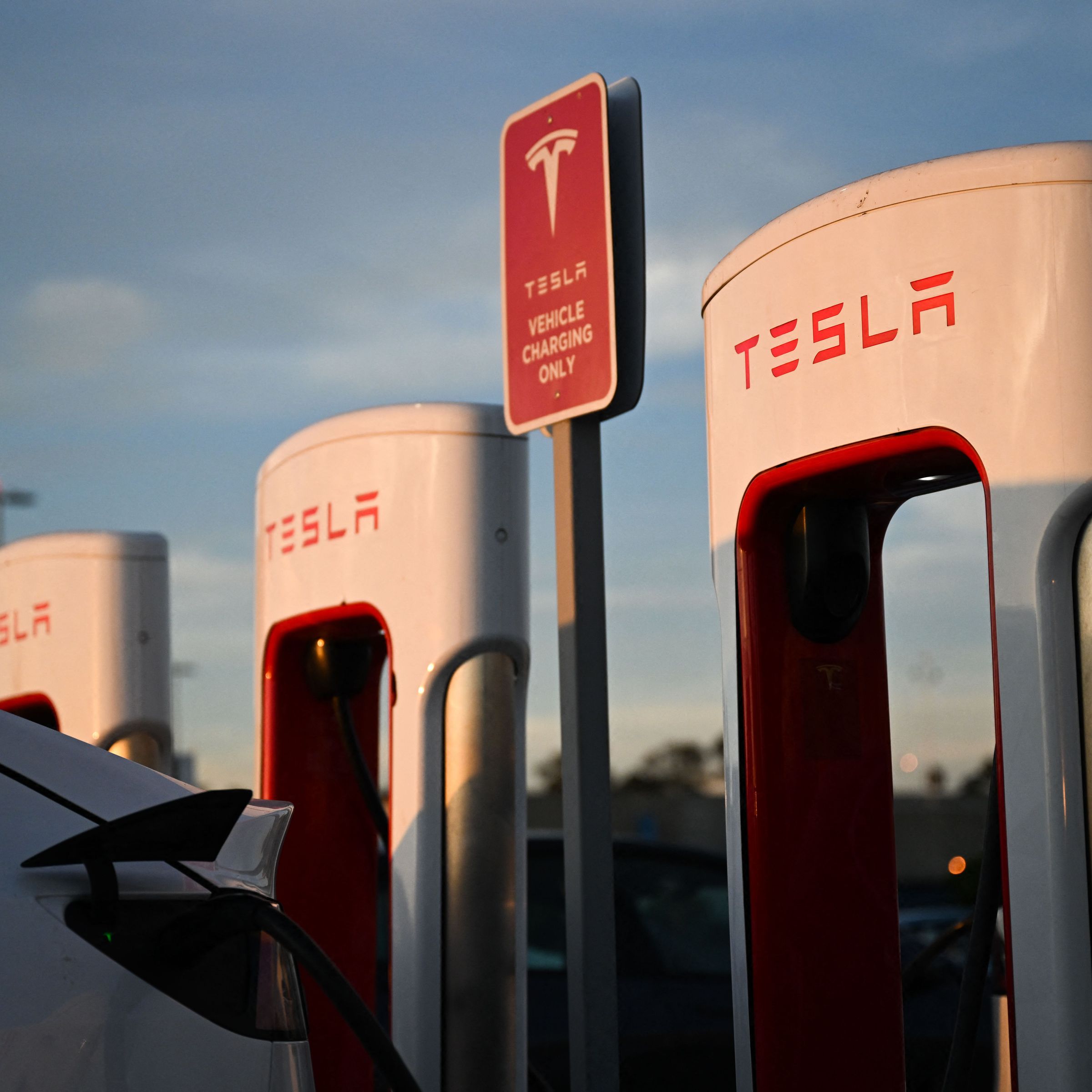 Tesla Supercharger station against a sunset and blue sky, and a Model Y is charging at one of the three stations in the shot