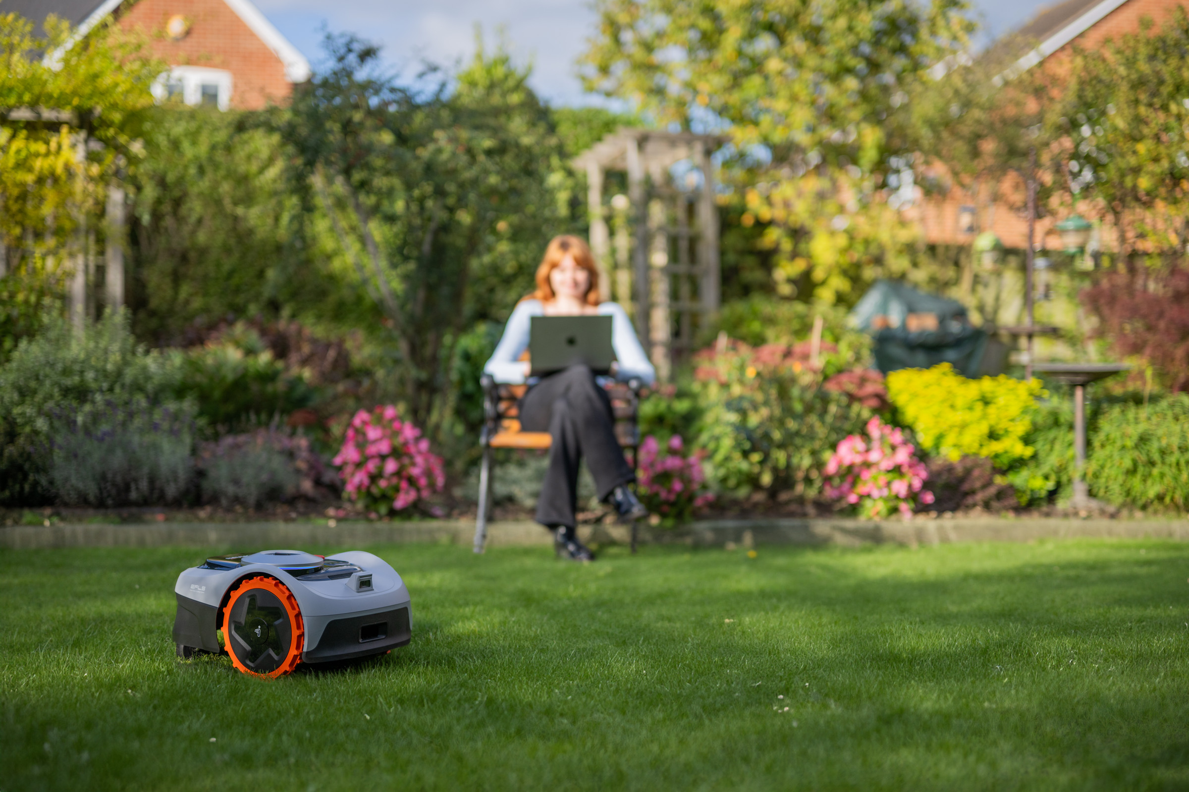 A robot lawnmower on a lawn with a lady sitting in a chair on her computer.