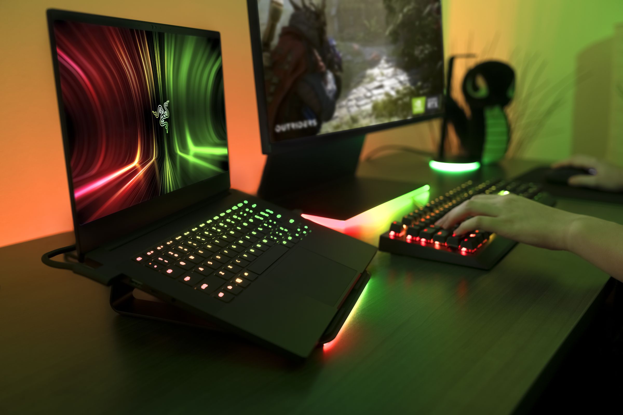 A user uses the Razer Blade 14 on a laptop stand connected to an external mechanical keyboard and monitor. The monitor screen displays a game; the laptop screen displays a Razer logo on a red, black, and green background.