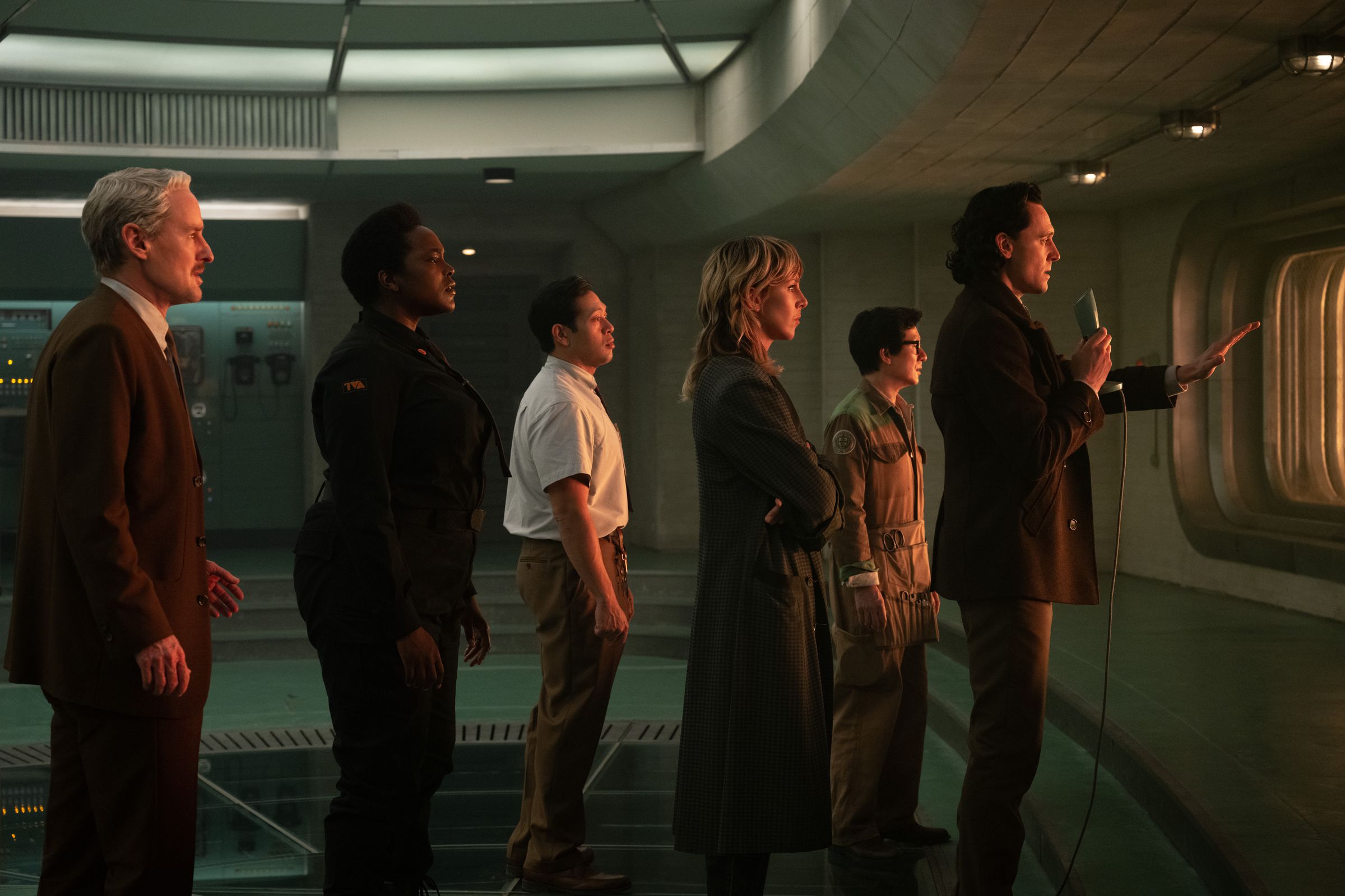 A man in a suit, a woman in a futuristic security uniform, a man in a short sleeved shirt and slacks, a woman in a long coat, a man in a gray jump suit, and a man in a tweed coat in slacks holding a microphone as they all gaze out of a large window in what looks like a space ship.