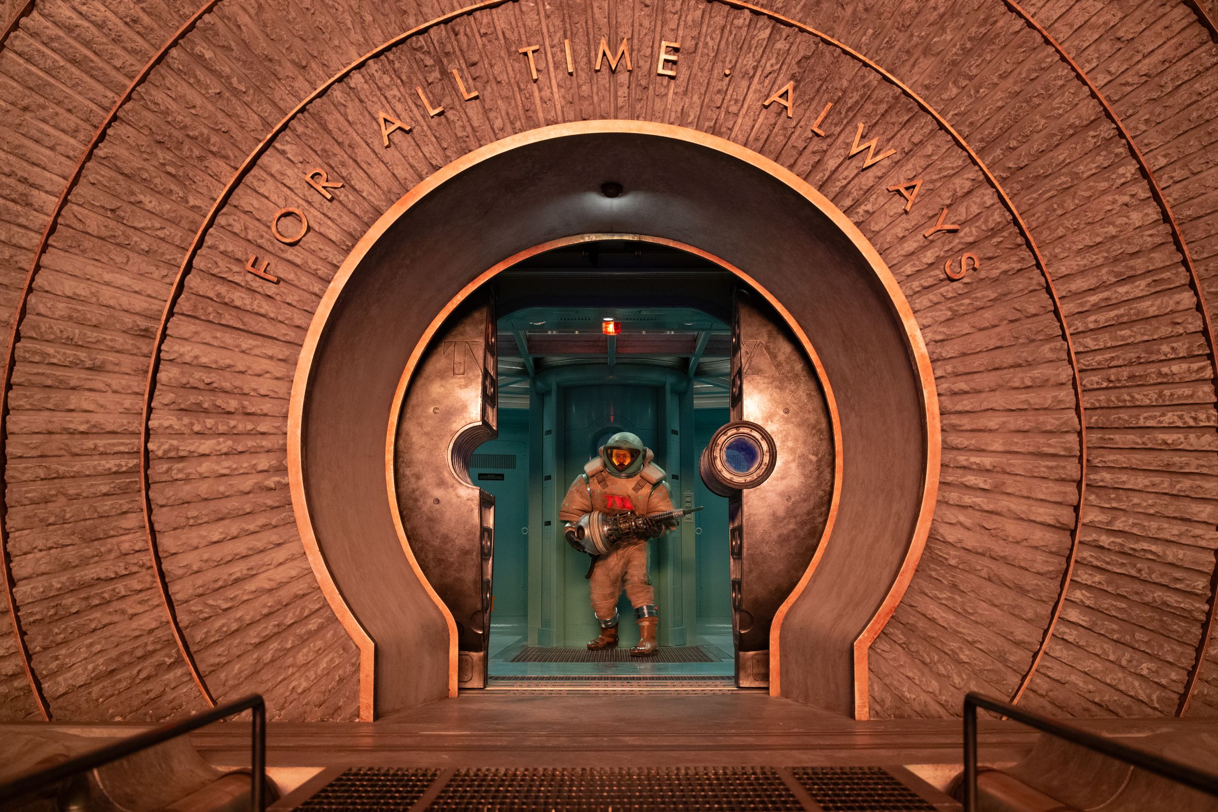 A man in a spacesuit holding a device that looks like a massive screw in both hands. The man is standing in the circular doorway opening onto a platform with a grated gangway. Above the door the words “for all time. Always” are inscribed.