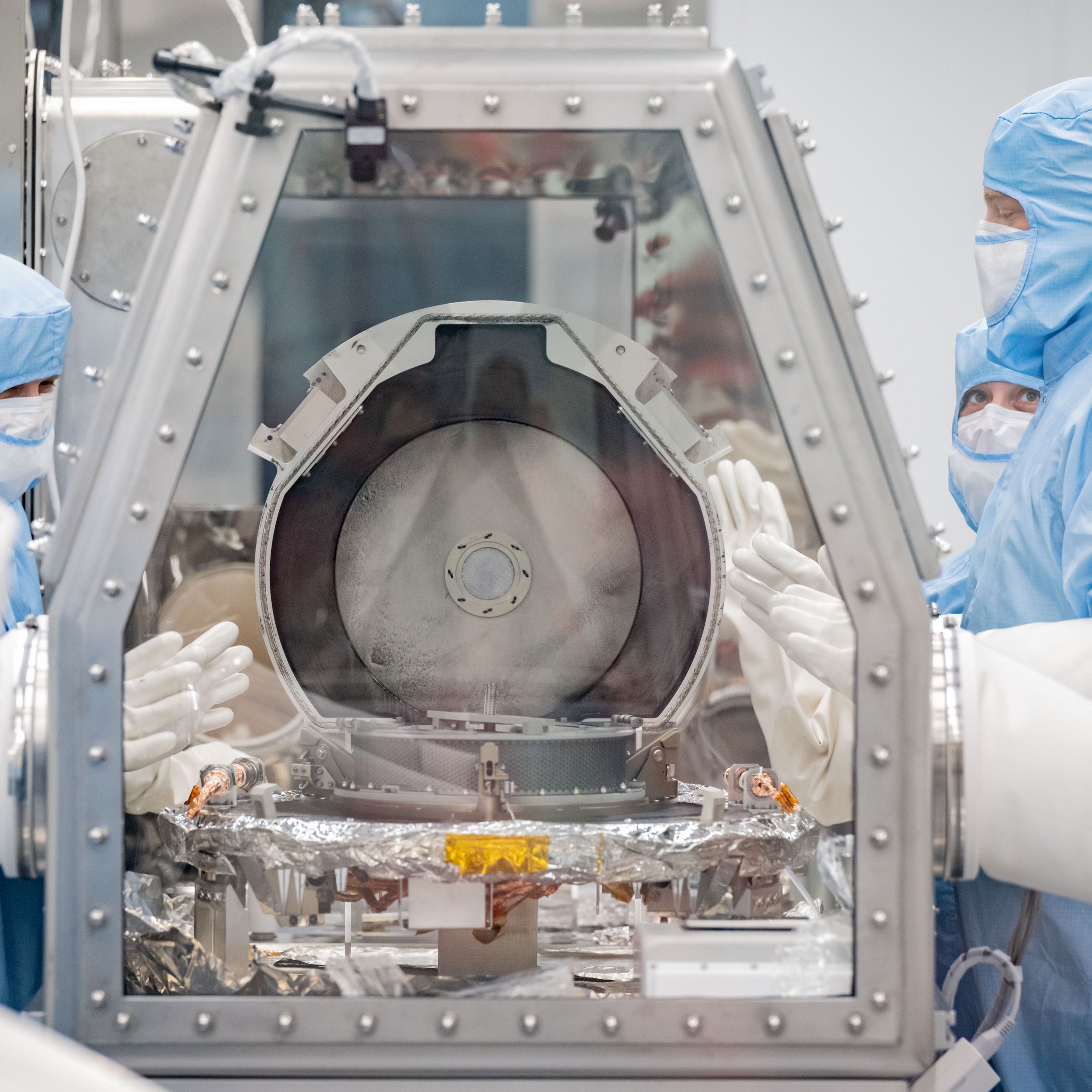 Several scientists in a clean room wearing protective gear look into the glass-shielded container holding the OSIRIS-REx sample return canister.