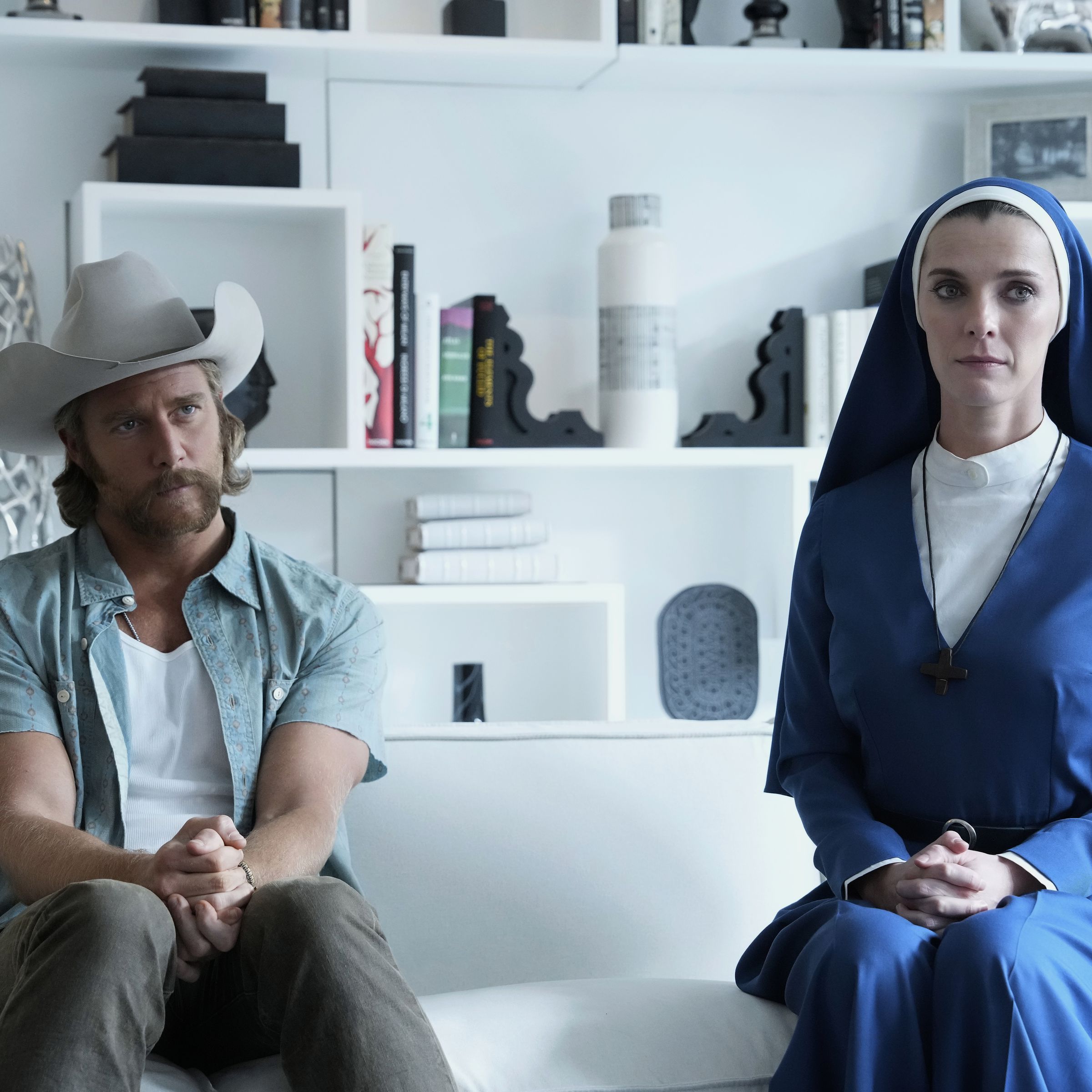 A white man wearing a white cowboy hat and sporting a very large blond mustache sits next to a white woman in a blue nun habit. They are seated very far apart on a white couch in a white room.