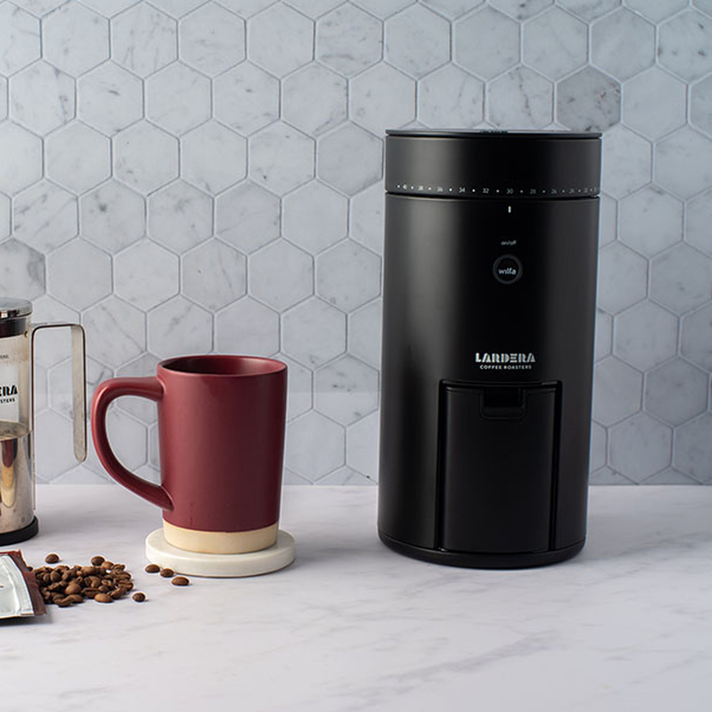 From left to right, a plant, a French press coffee maker, a reddish-brown mug, and a black, conical coffee grinder