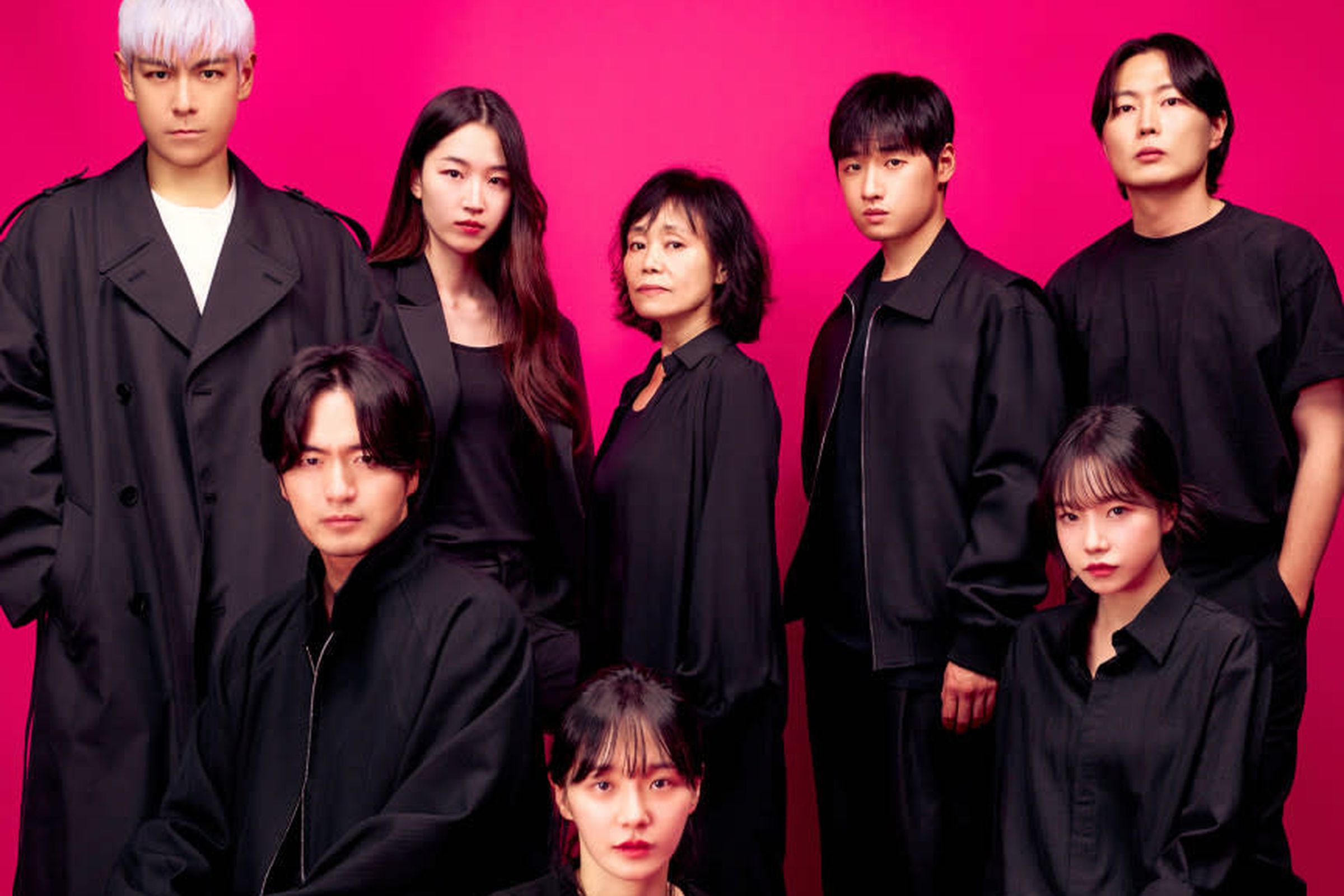 A group of eight men and women who are all wearing head-to-toe black outfits, and standing together in front of a bright pink wall.