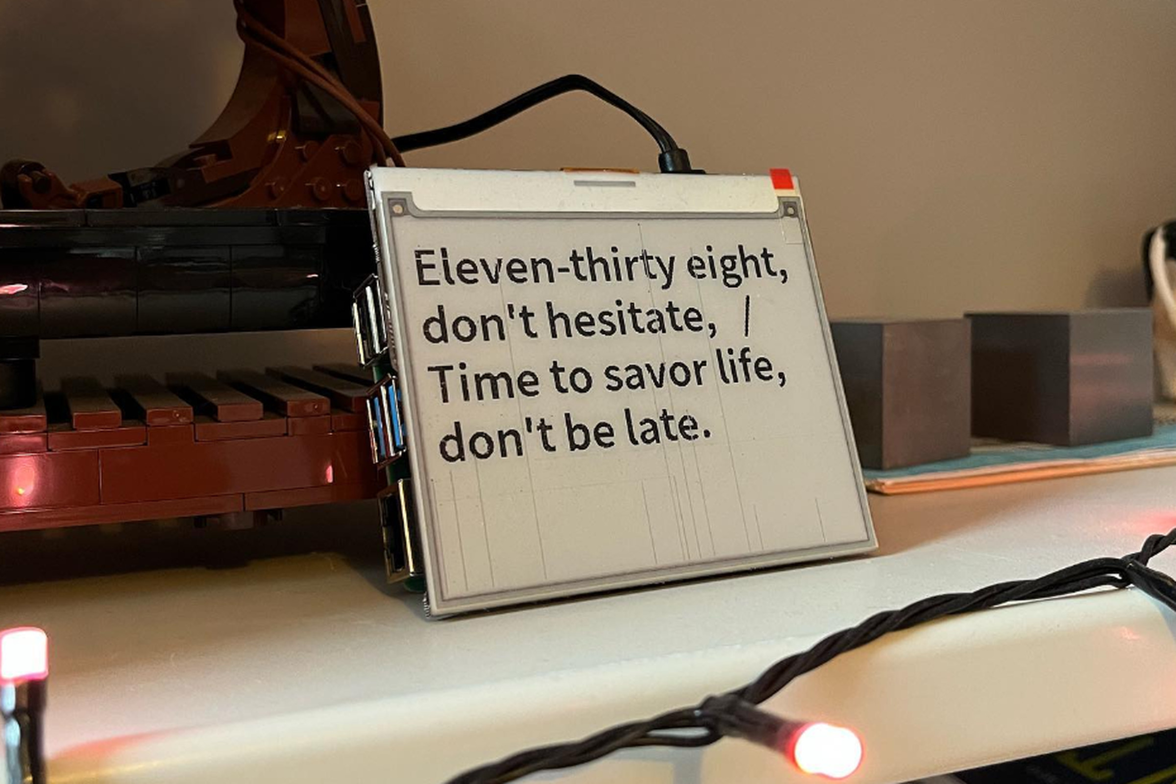 A photograph of a low-fi E Ink screen sitting on a bookshelf. The text on the clock says “Eleven-thirty eight, don’t hesitate, / Time to savor life, don’t be late.”