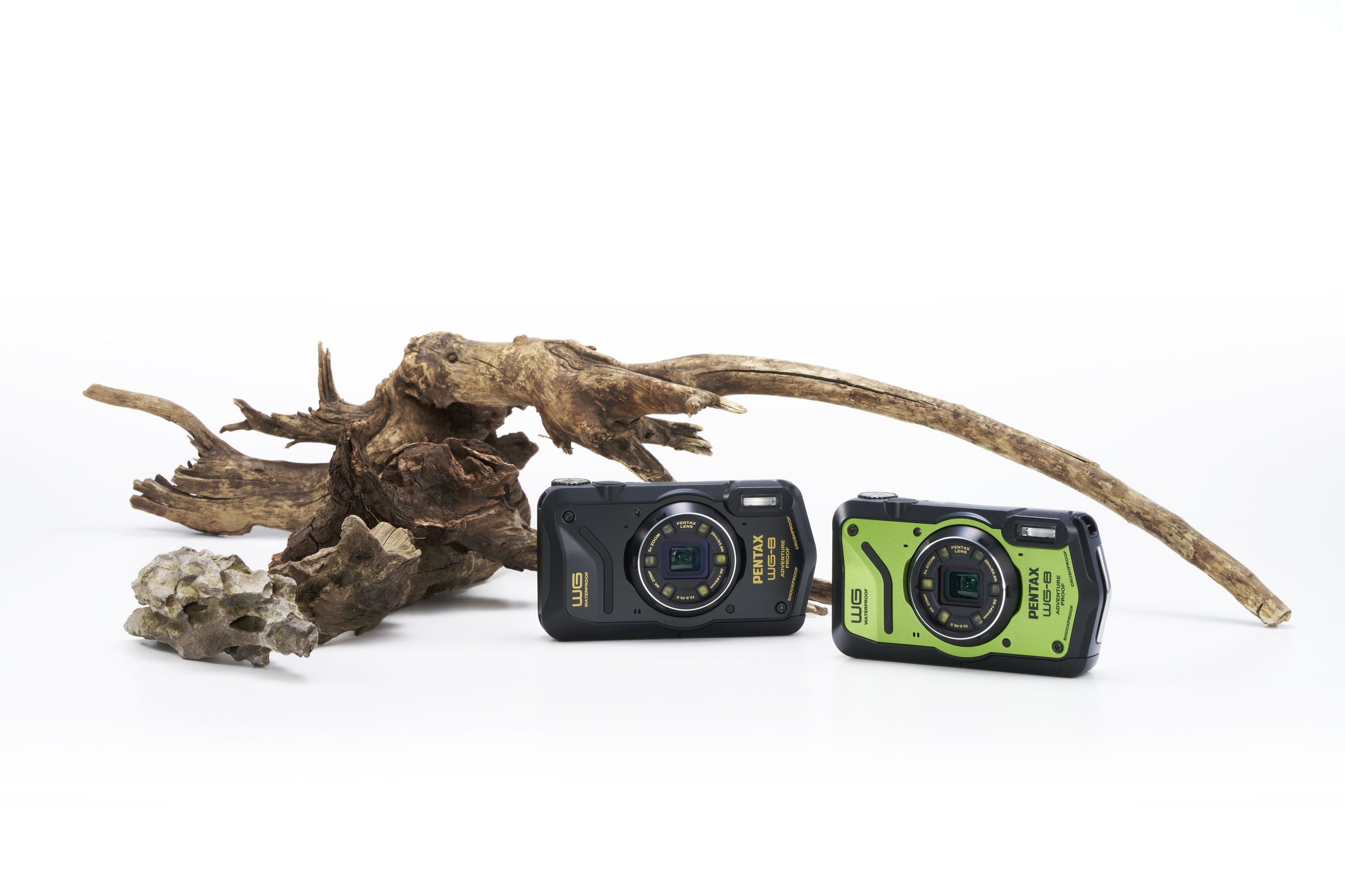 The Pentax WG-8 cameras in black and green next to some driftwood on a white background.