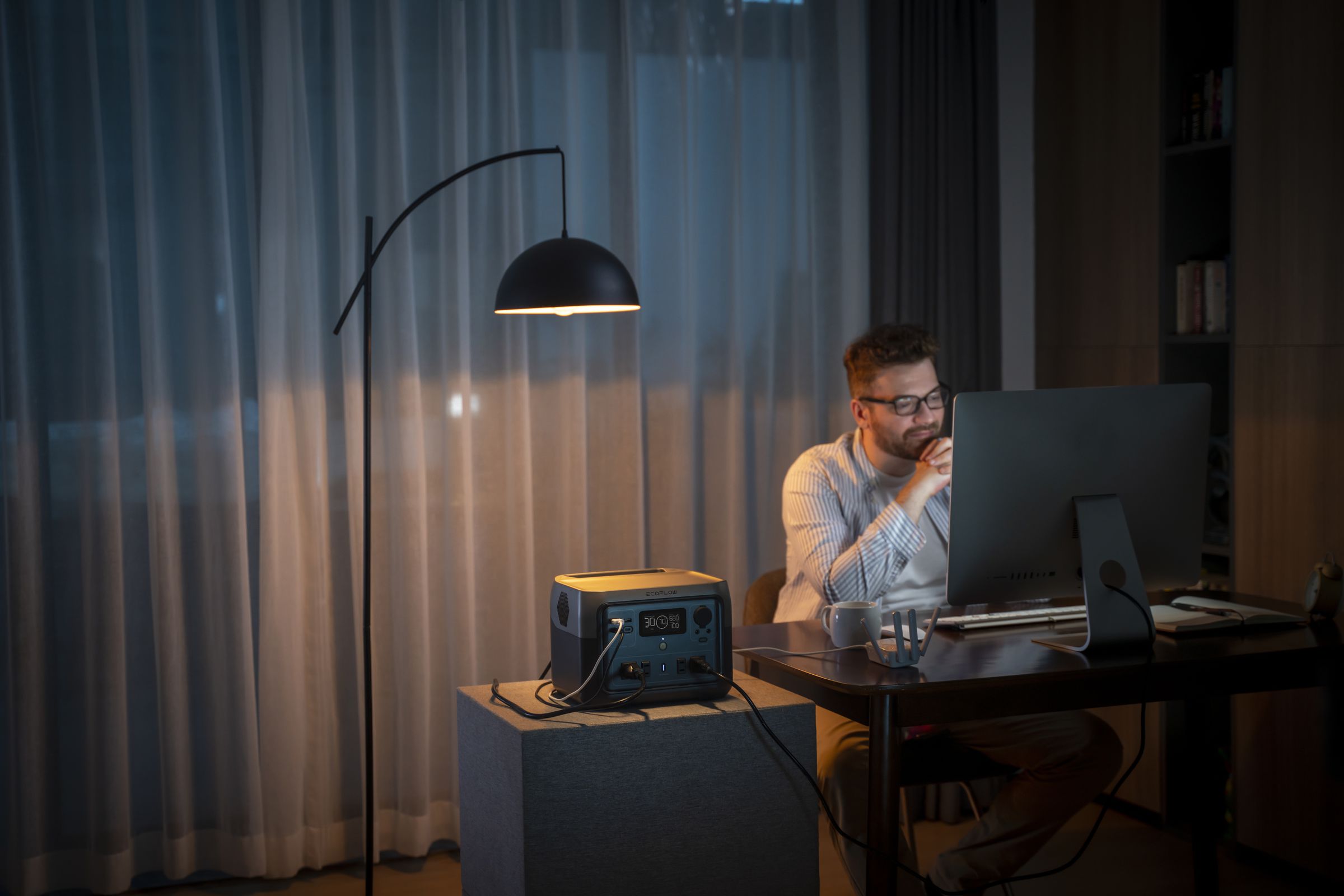 A River 2 Max battery powers a desktop computer and accessories while a man sits at his office under the light of a lamp.