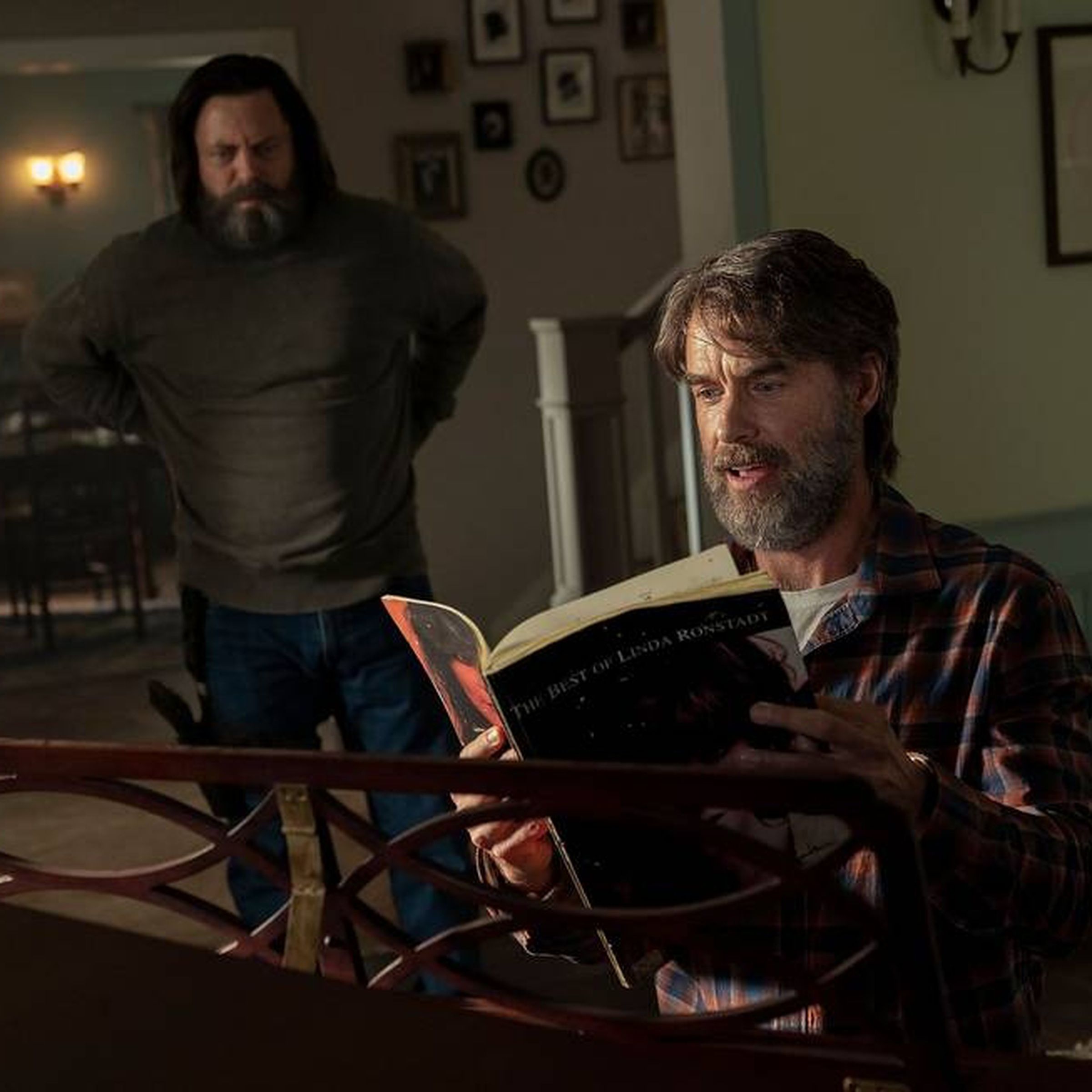 A bearded man wearing a flannel and holding a book of music while sitting at a piano. In the background, another bearded man wearing a sweater, and holding his hands on his hips is looking on.