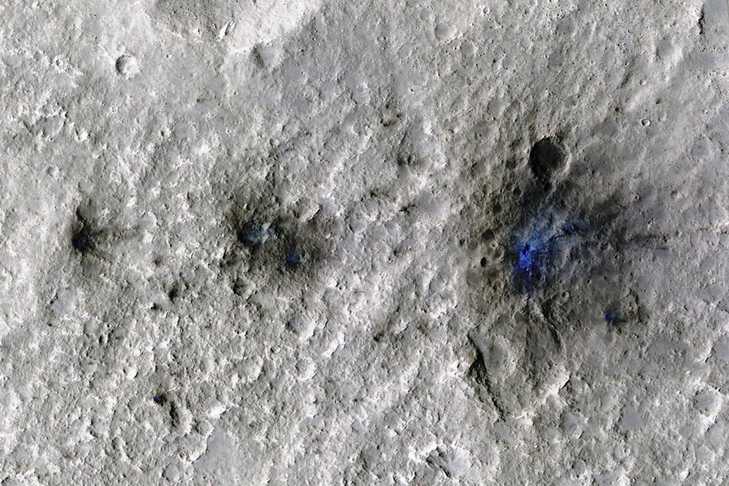 Three dark black craters on a pocked light grey surface. The smallest is on the left and the largest is on the right. There are faint traces of bright blue in the center of each.