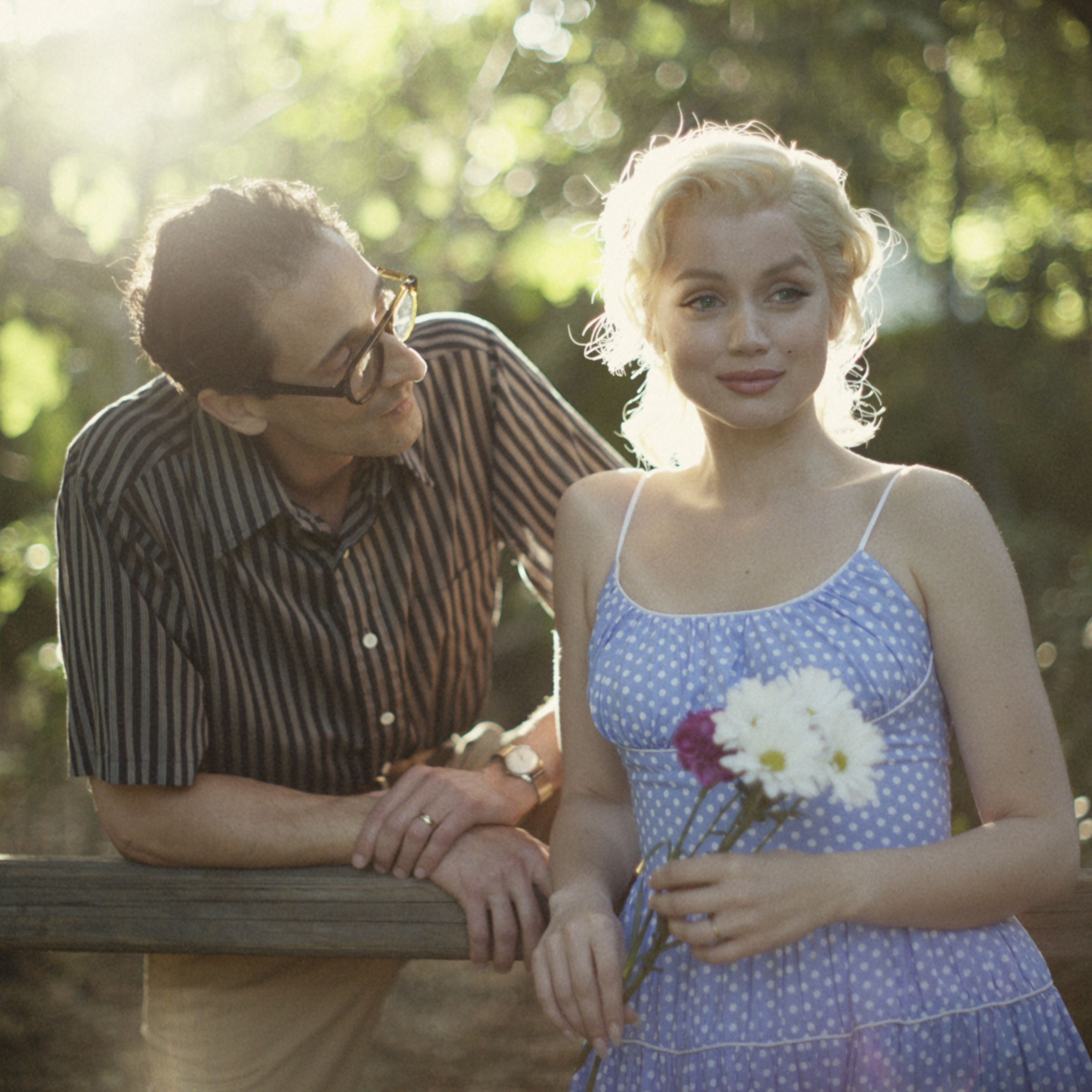 A bespectacled man leaning on a wooden fence as he gazes at a woman holding a bunch of flowers and smiling off into the distance.