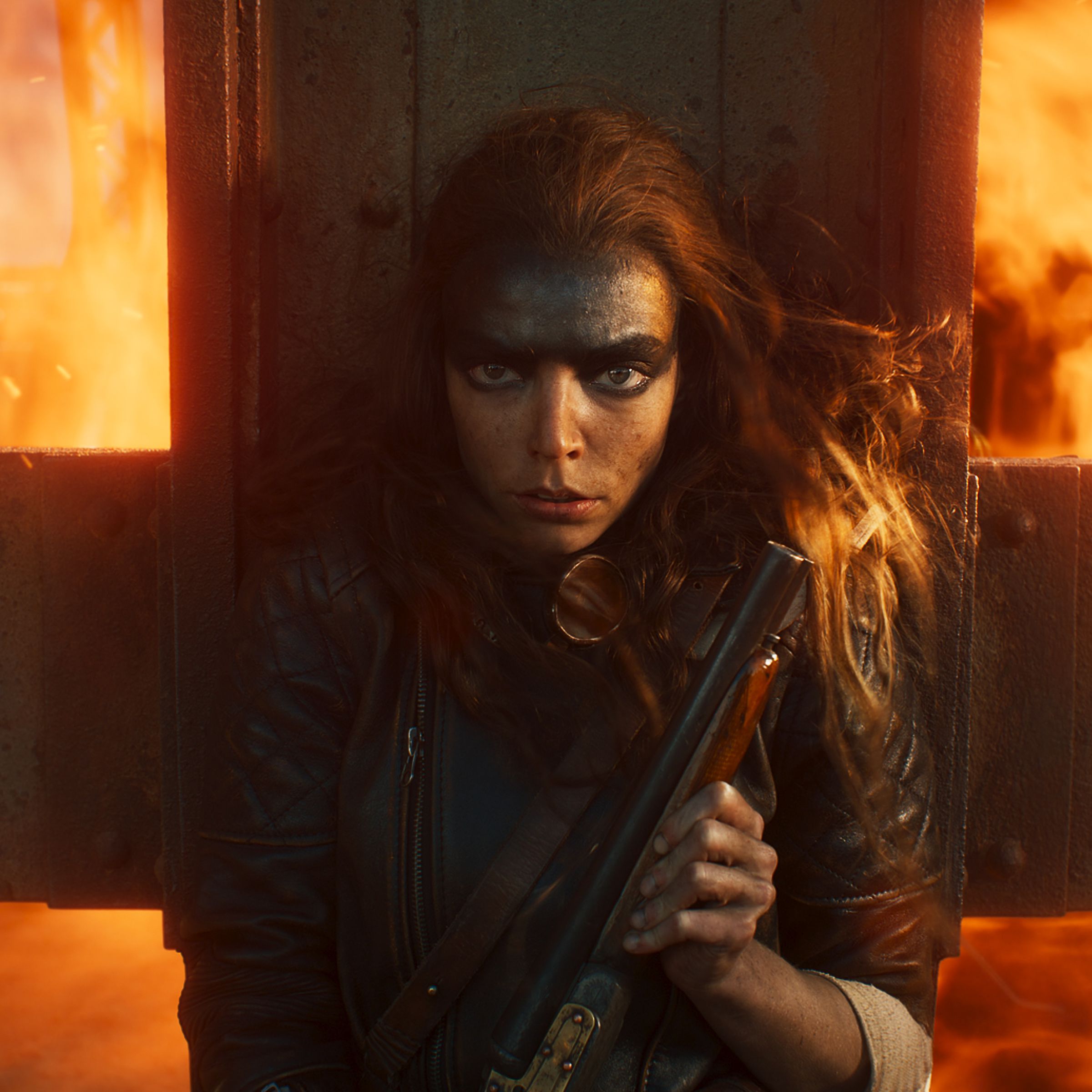A woman clutching a shotgun to her chest while pressing up against a metal grate to protect herself from a wall of flames rushing up from behind her.