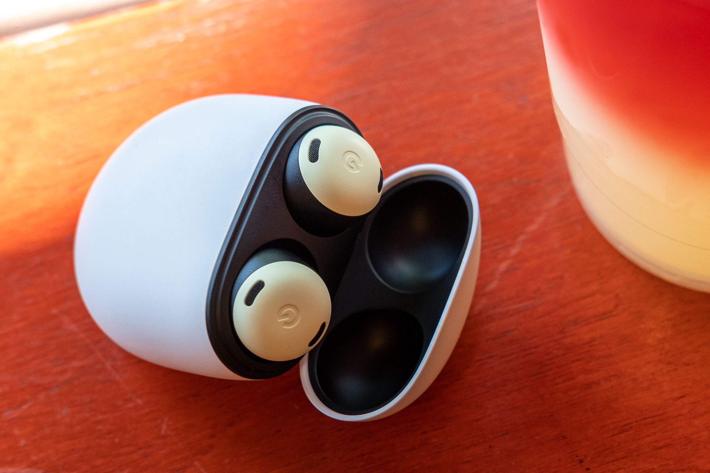 The yellow Pixel Buds Pro wireless earbuds sitting in their charging case with the lid open, resting on an orange table beside a plastic cup of colorful lemonade.