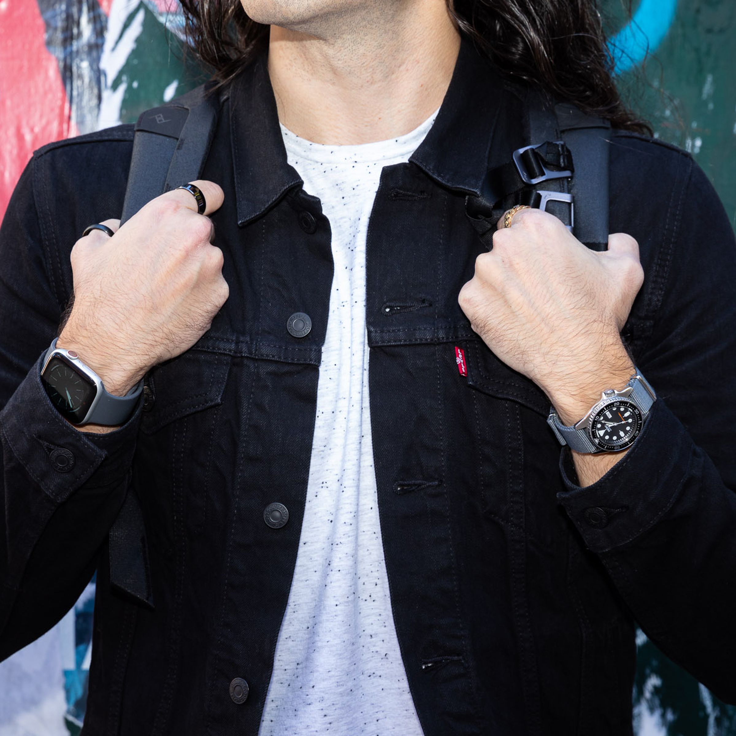 A close-up photo of a man wearing a black denim jacket, holding his backpack straps and exposing both a traditional watch and an Apple Watch on his wrists.
