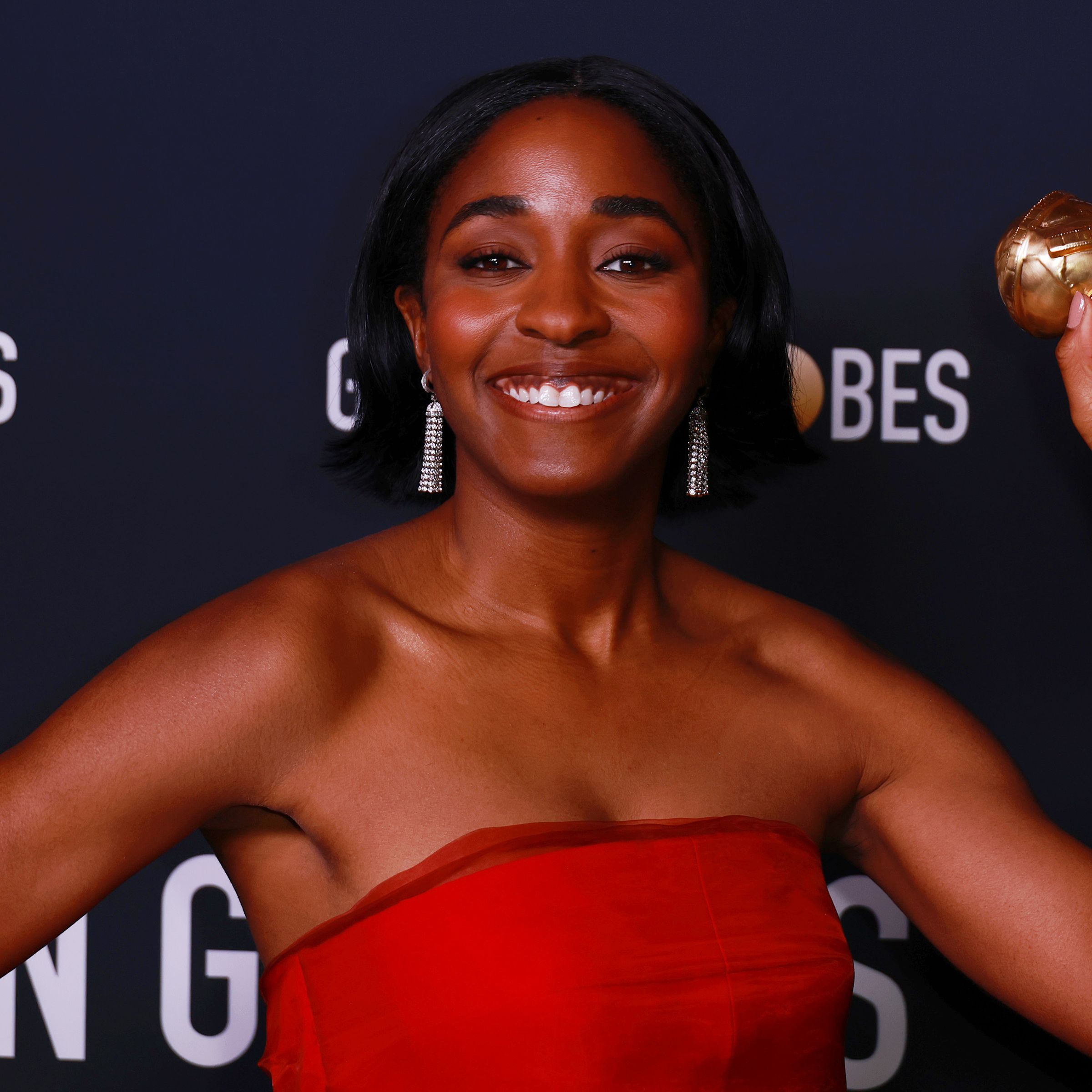 A Black woman with a bob smiling, wearing a strapless red dress and clutching a small golden statue topped with a globe in her upraised right arm.