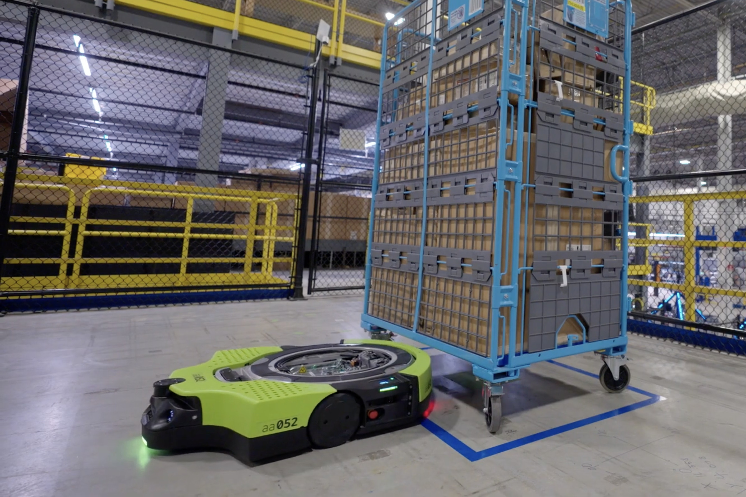 Image of a green robot sliding out from under a big metal cart filled with boxes.