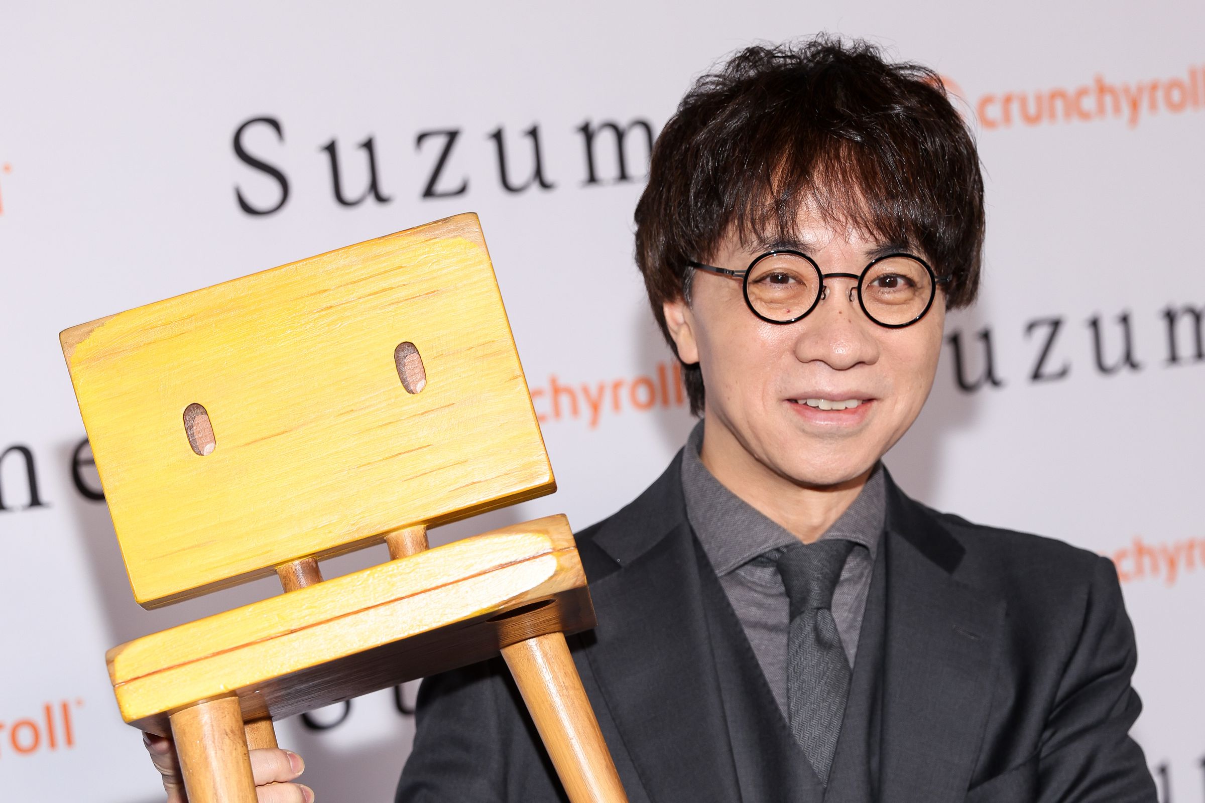 A bespectacled man in a black suit with a steel gray shirt and tie holding up a wooden chair.