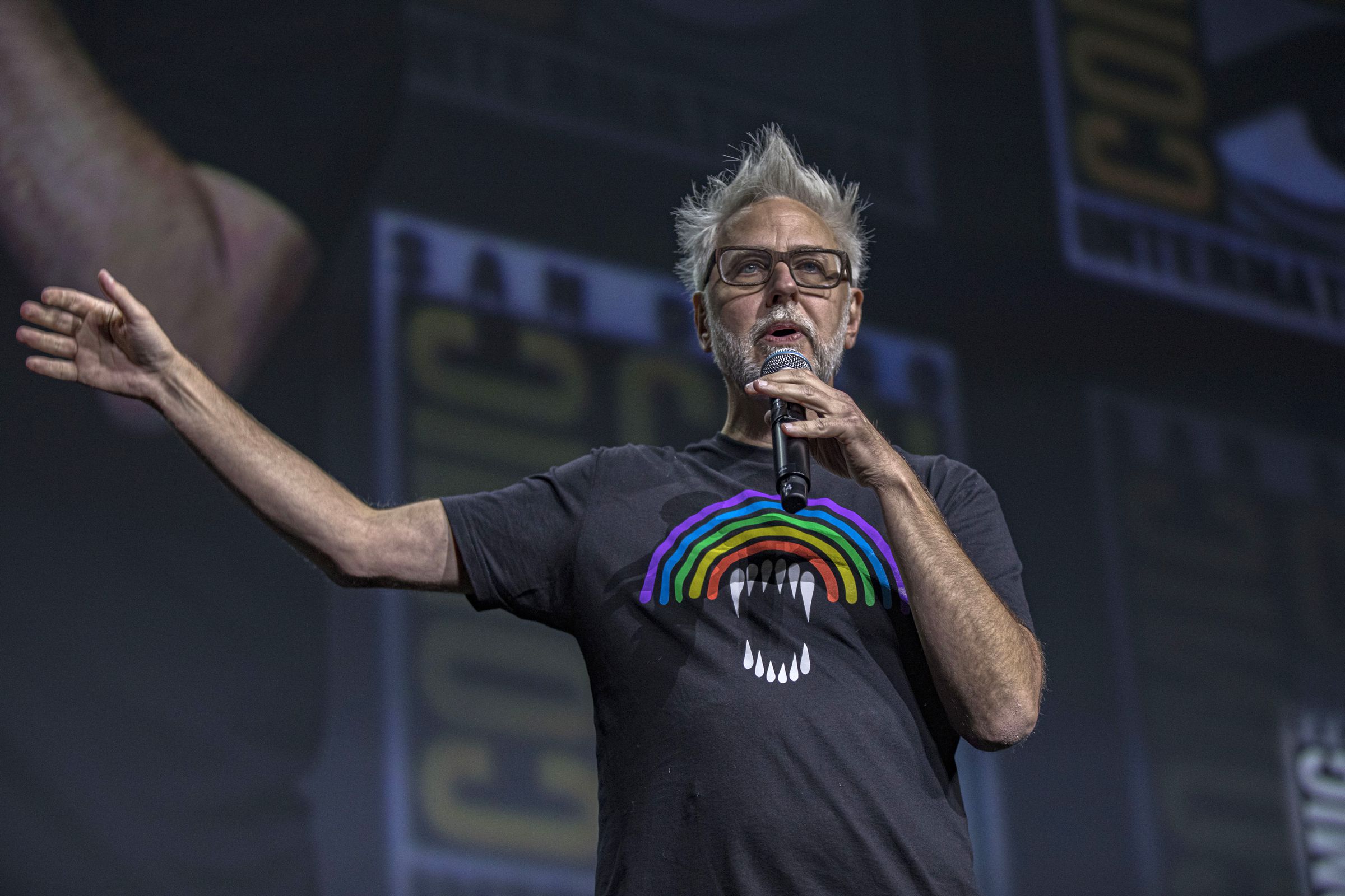 James Gunn wearing a black t-shirt with a rainbow on it as he gestures on stage during a presentation at San Diego Comic-Con in 2022.