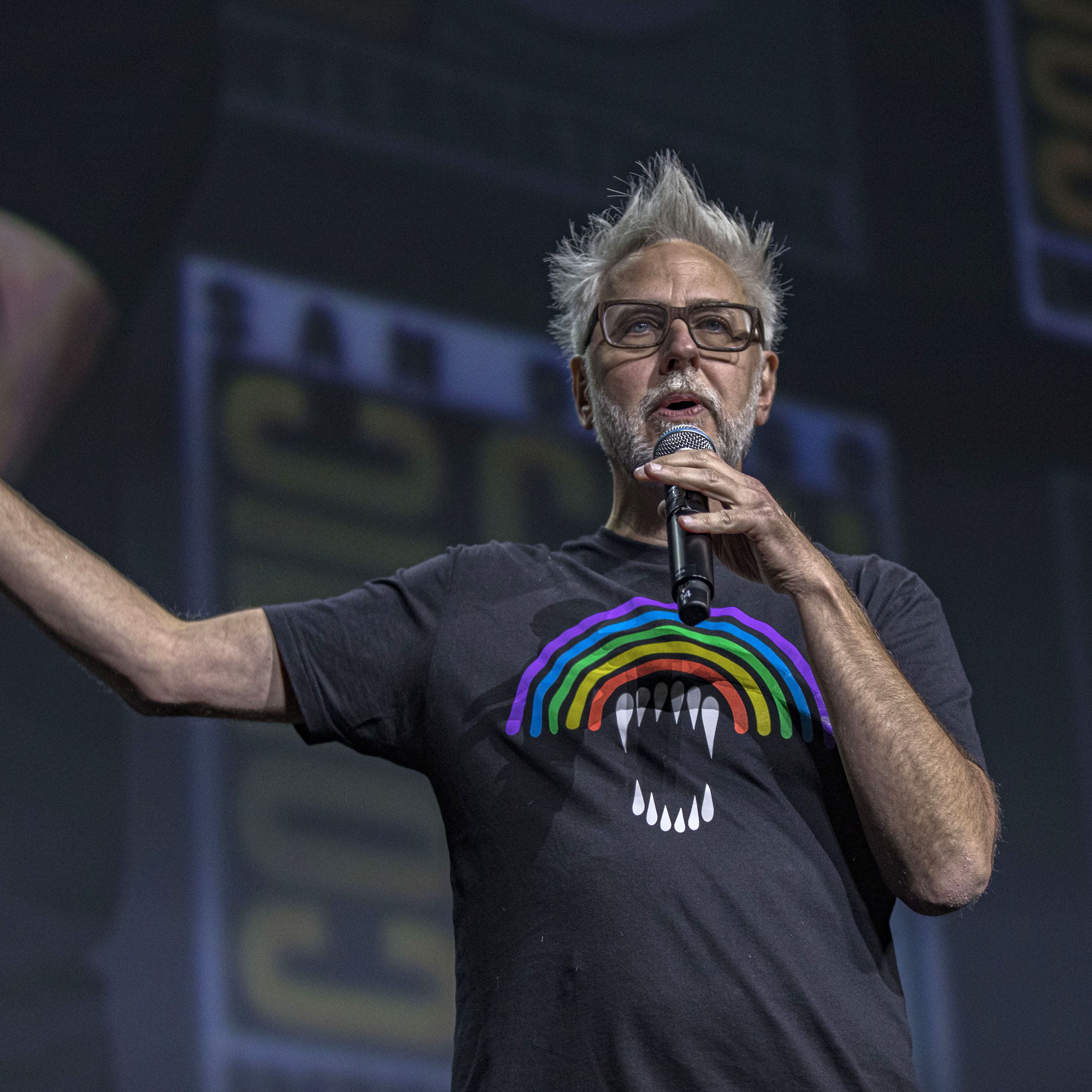James Gunn wearing a black t-shirt with a rainbow on it as he gestures on stage during a presentation at San Diego Comic-Con in 2022.