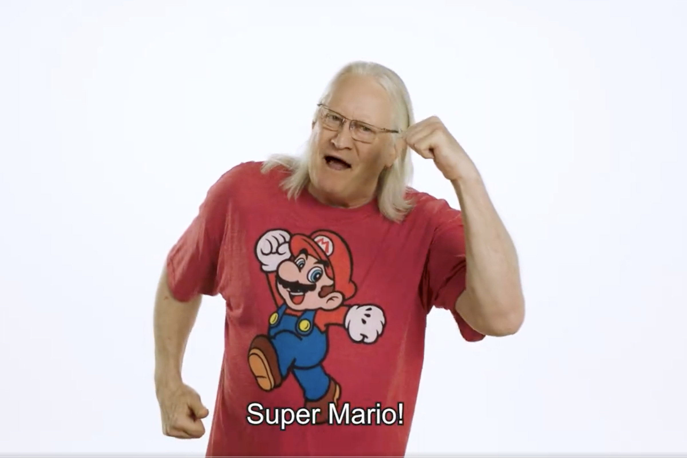 Screenshot from a Nintendo video featuring Charles Martinet, an older Caucasian man wearing a red Mario shirt, holding up his arms in a running motion saying, “Super Mario.”