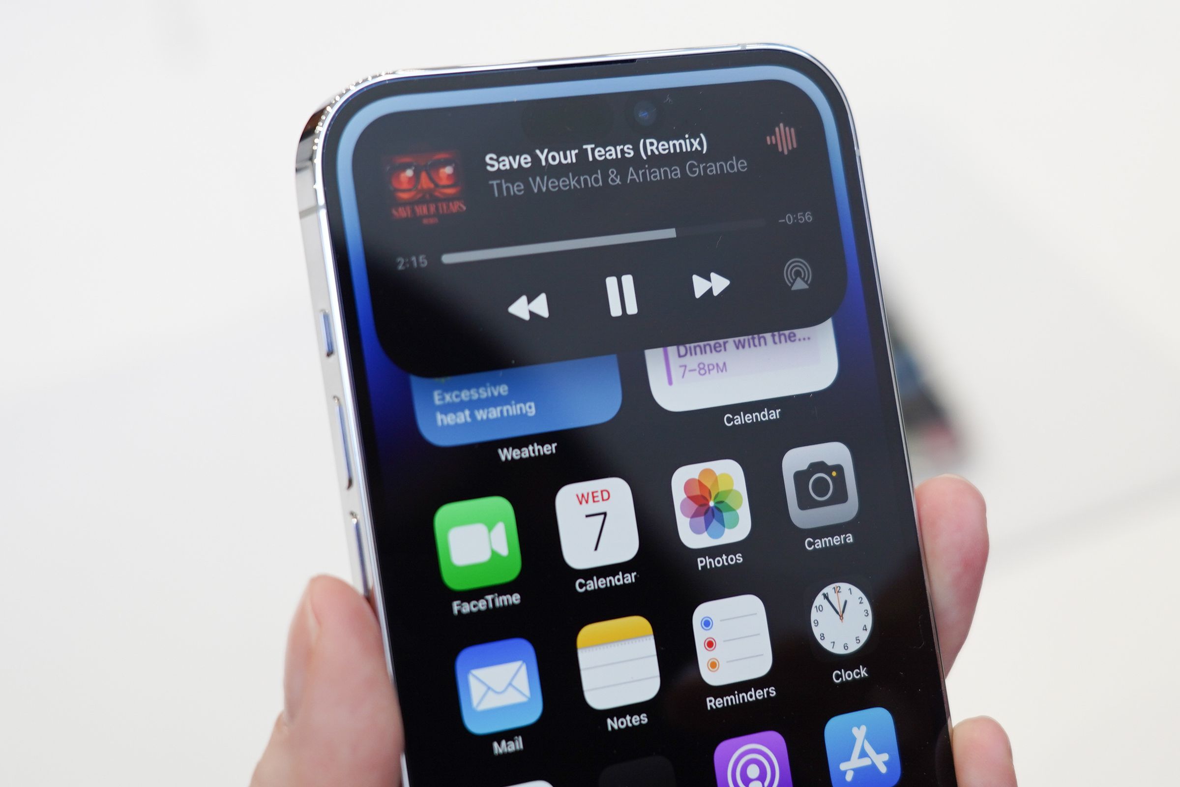 An image of an iPhone held in a hand. The Dynamic Island feature is prominent on the display. It’s a black box that shows a music player in it.
