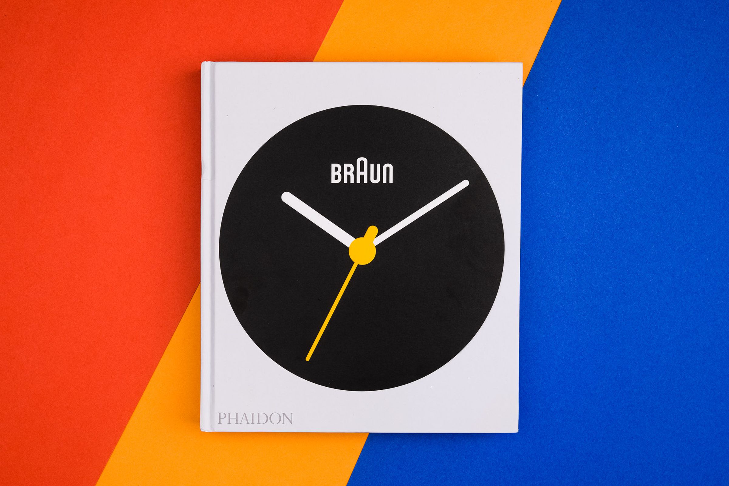 Photo of the cover of the book Braun: Designed to Keep, which features a large black Braun watchface with orange minute hand on white background. The book sits flat on a diagonally striped red, orange and blue table.