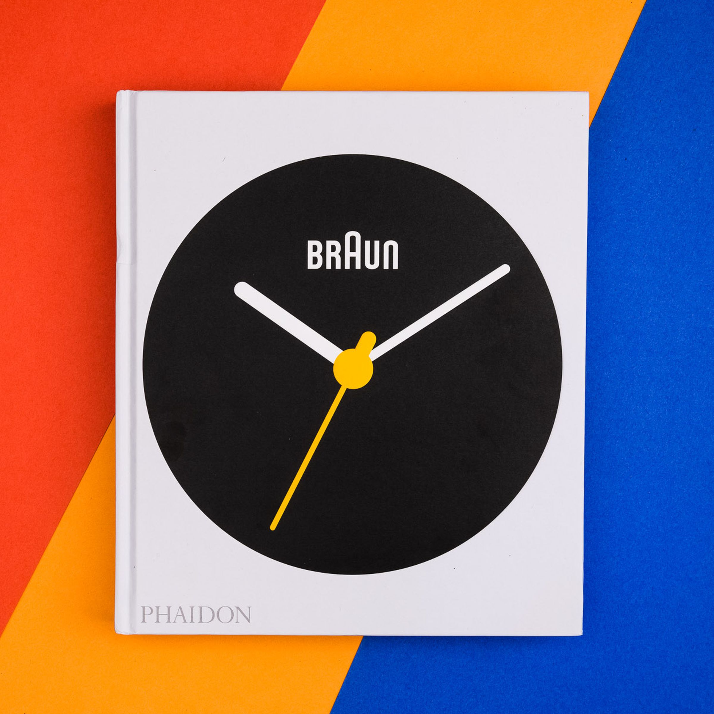 Photo of the cover of the book Braun: Designed to Keep, which features a large black Braun watchface with orange minute hand on white background. The book sits flat on a diagonally striped red, orange and blue table.