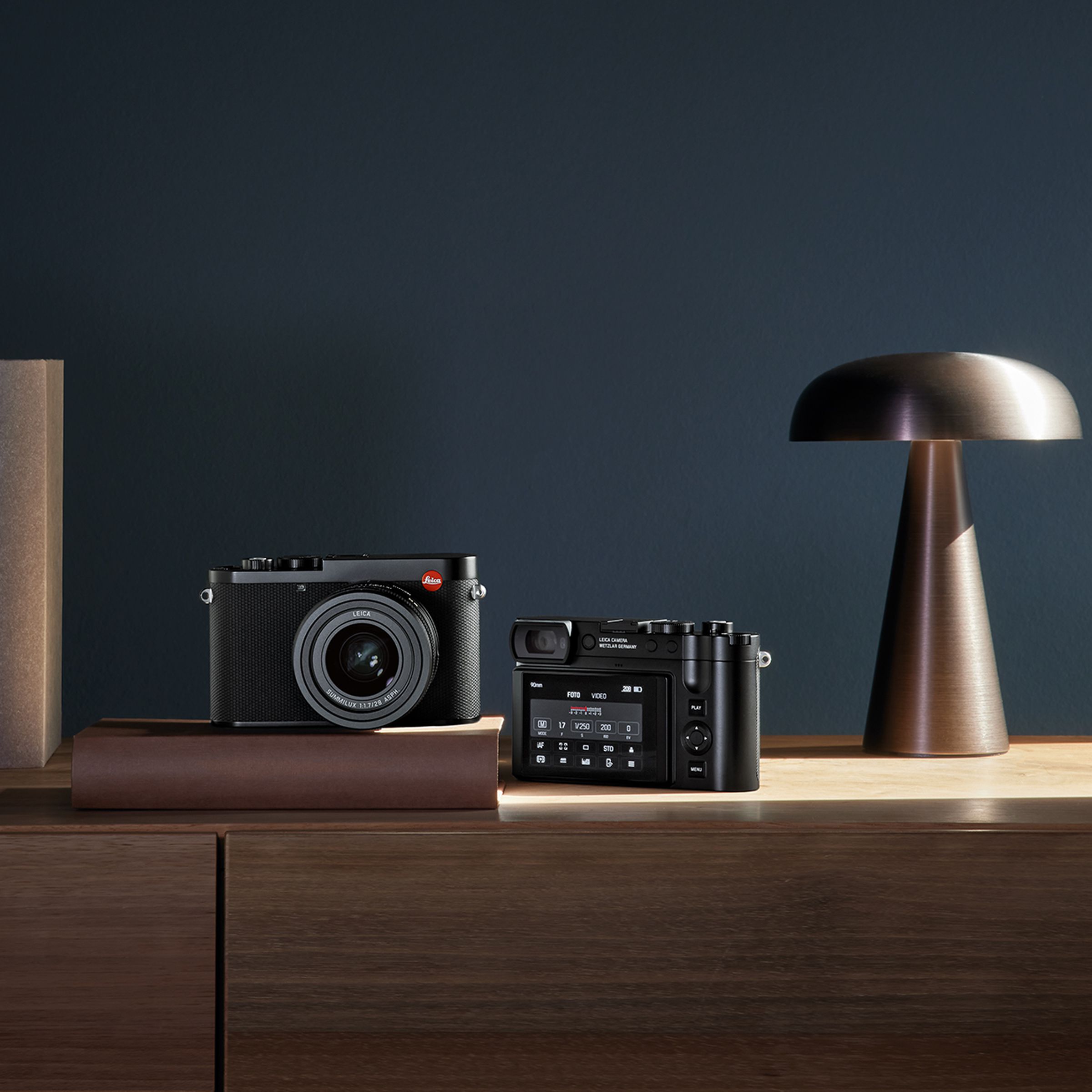 Two Leica Q3 cameras sitting on a piece of wood furniture, one facing forward and one facing away.