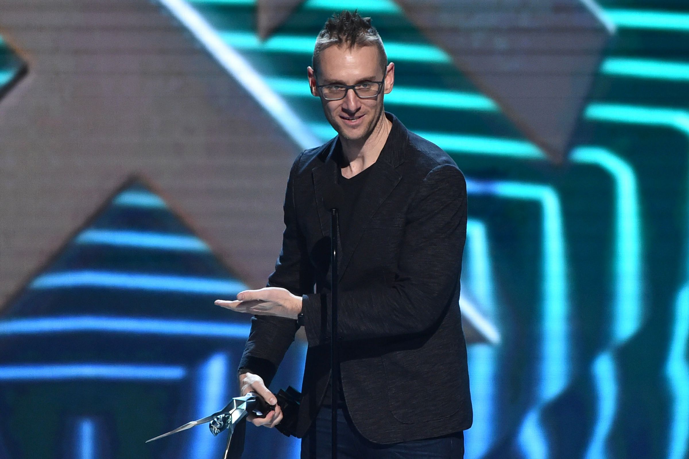 A man with glasses and a sharp nose holds an awards statue onstage