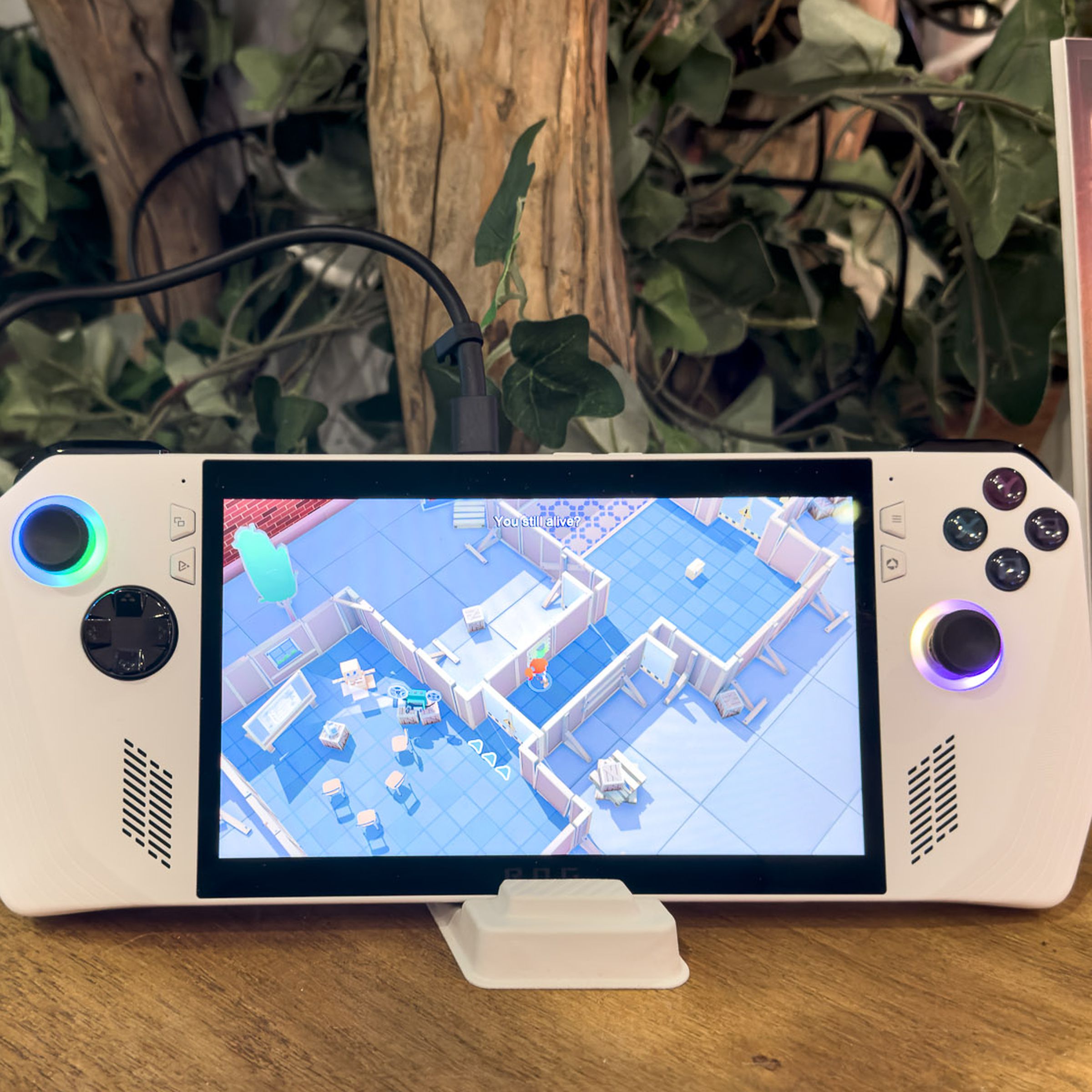 A white portable gaming handheld, a bit like a Nintendo Switch but with fixed controllers, on a stand on a wooden table with greenery in the background.