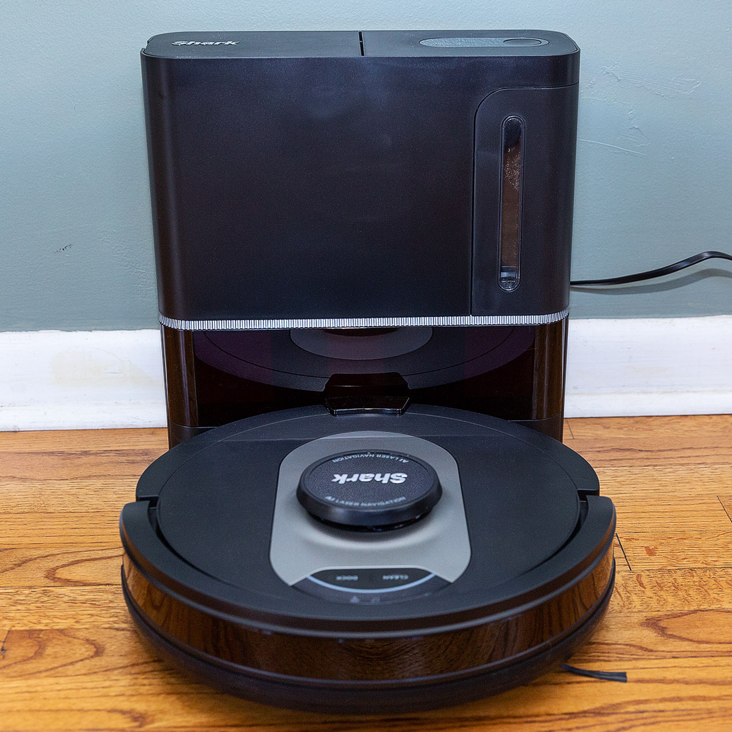 A head-on angle of the Shark AV2501AE AI Robot Vacuum docked in its self-cleaning station against a wall, plugged into a nearby outlet.